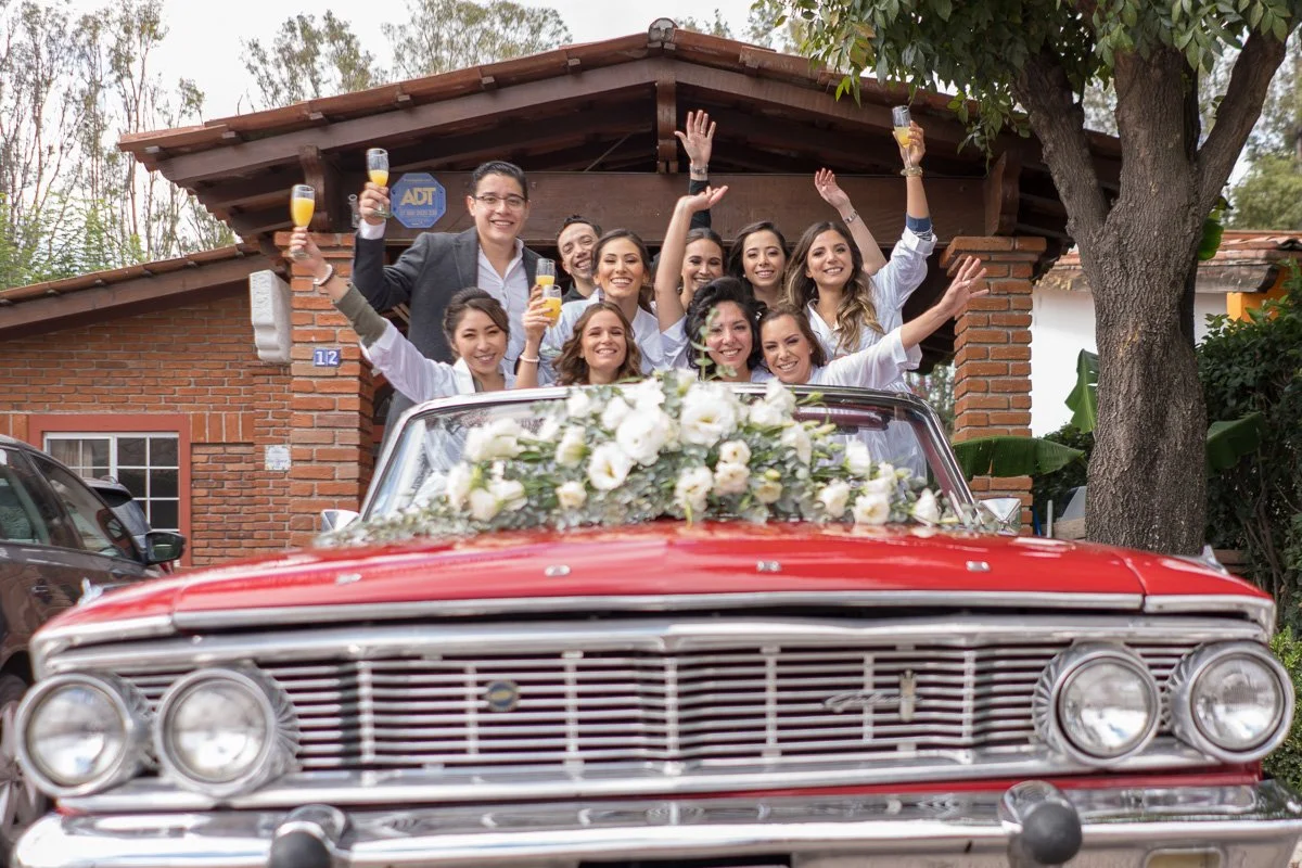Group of people celebrating on a decorated vintage red car with white flowers, holding glasses of champagne, in front of a brick house.
