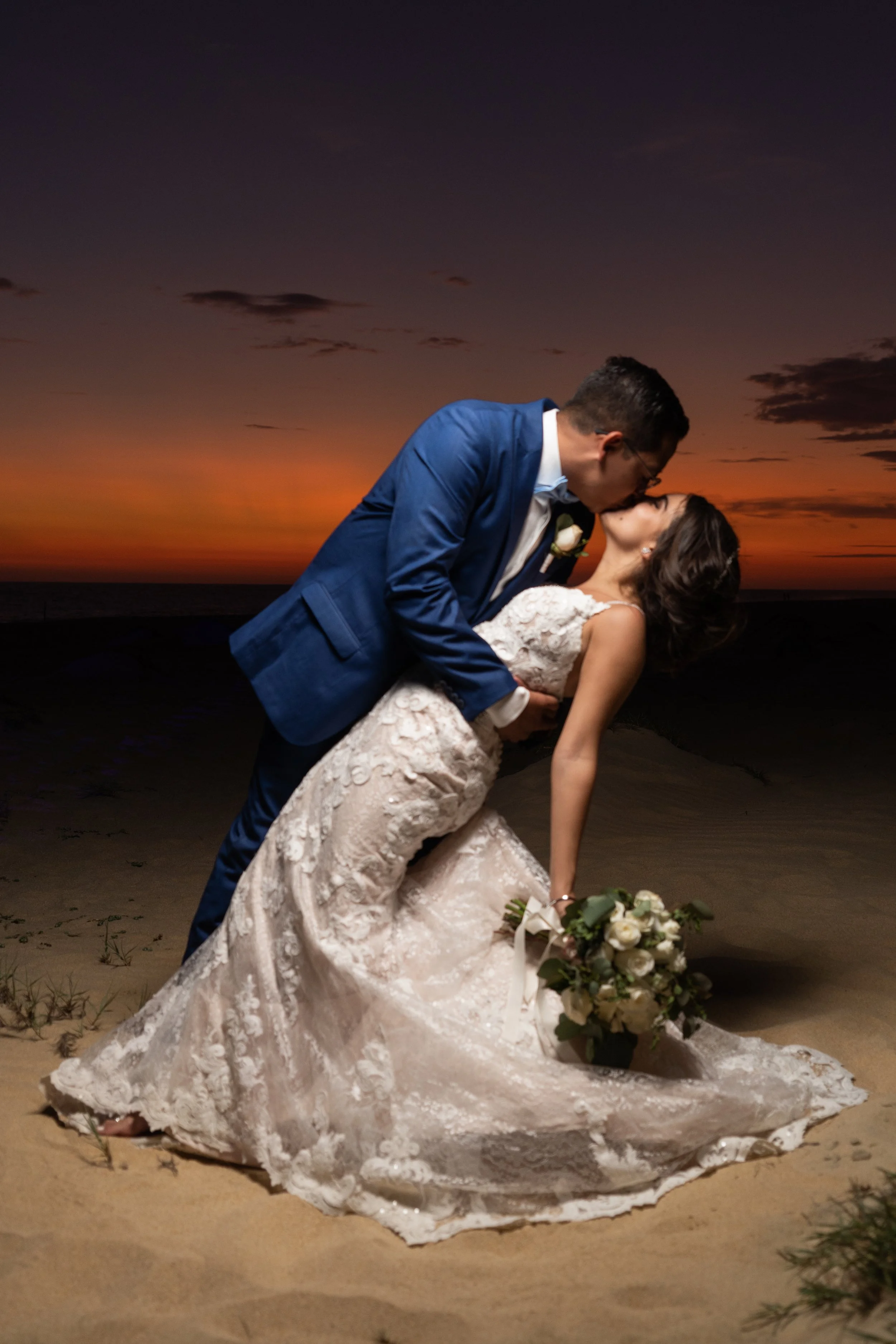 Pareja de novios en la playa al atardecer, la mujer con vestido de novia y ramo, el hombre con traje azul, en un beso romántico.