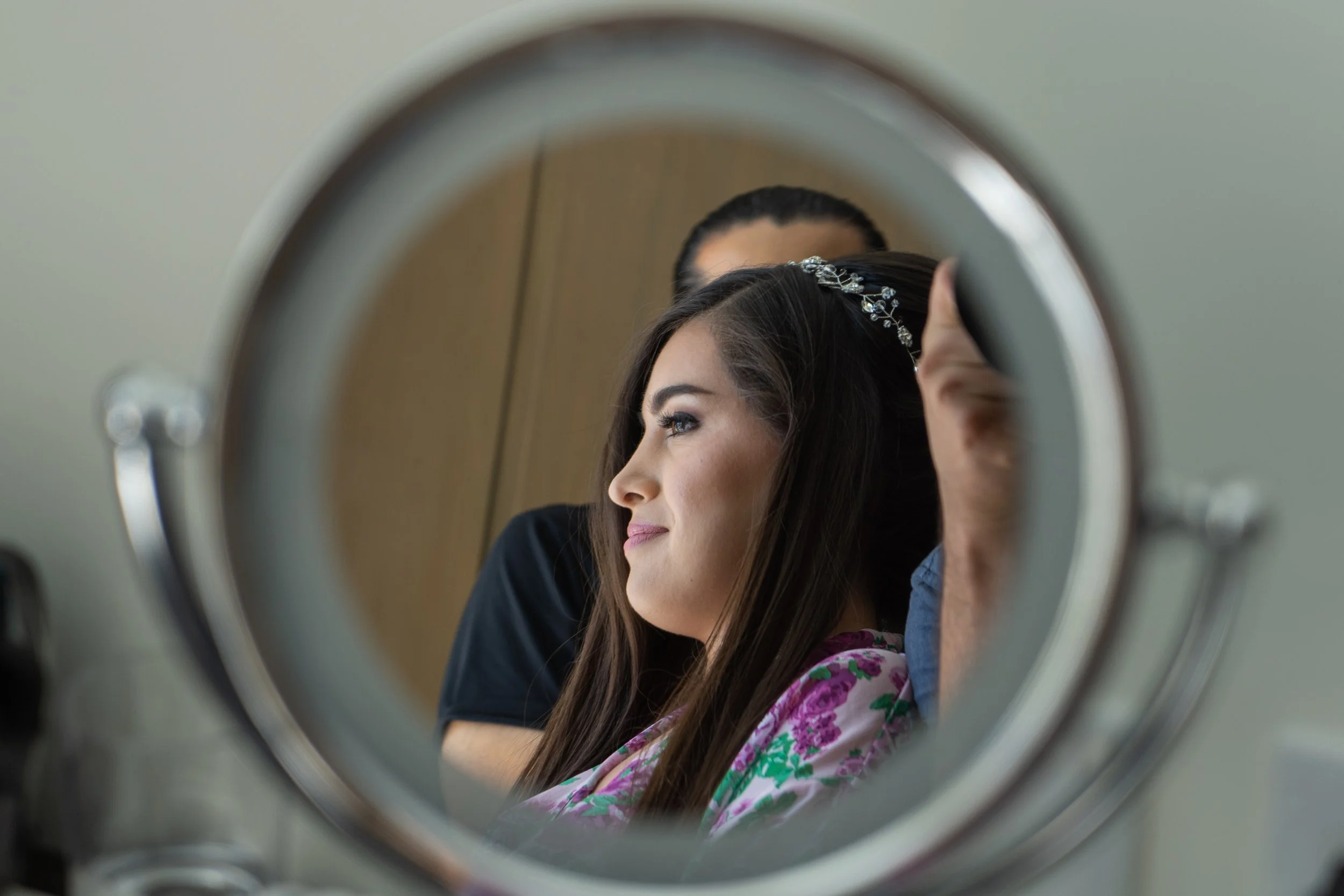 Mujer con peinado adornado con accesorios, viendo su reflejo en un espejo de tocador, mientras un ayudante la arregla en un cuarto con pared de color claro.