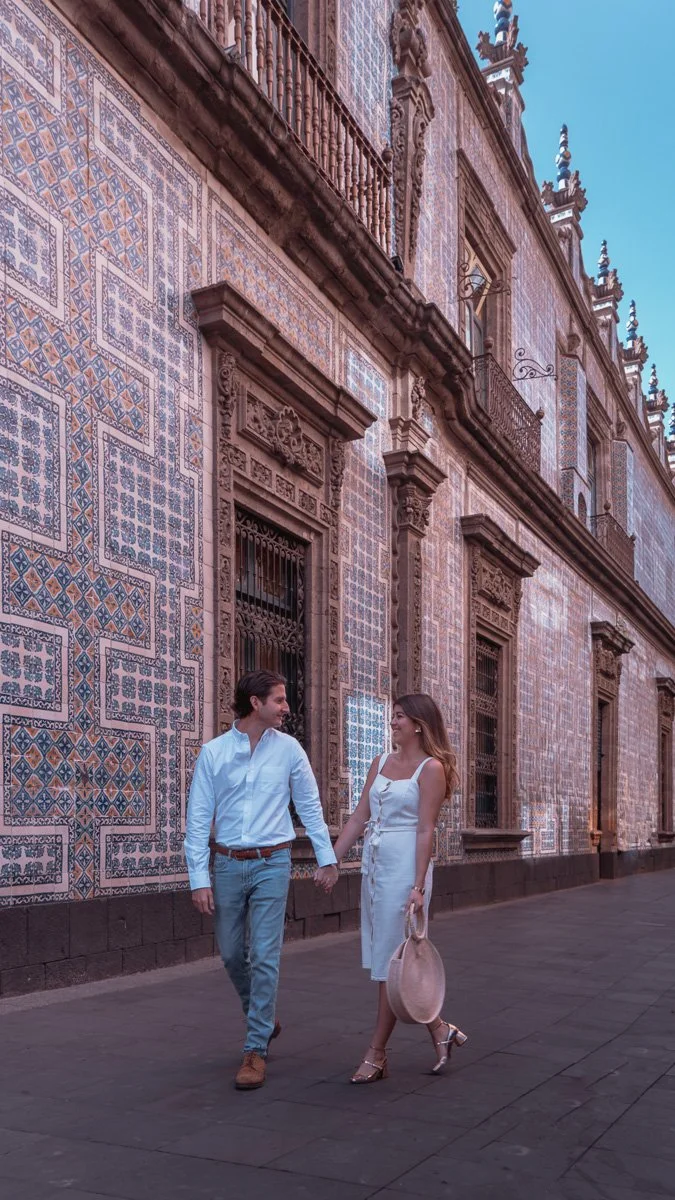 A couple walking hand-in-hand on a city sidewalk in front of a historic building with decorative tile work and ornate architectural details.