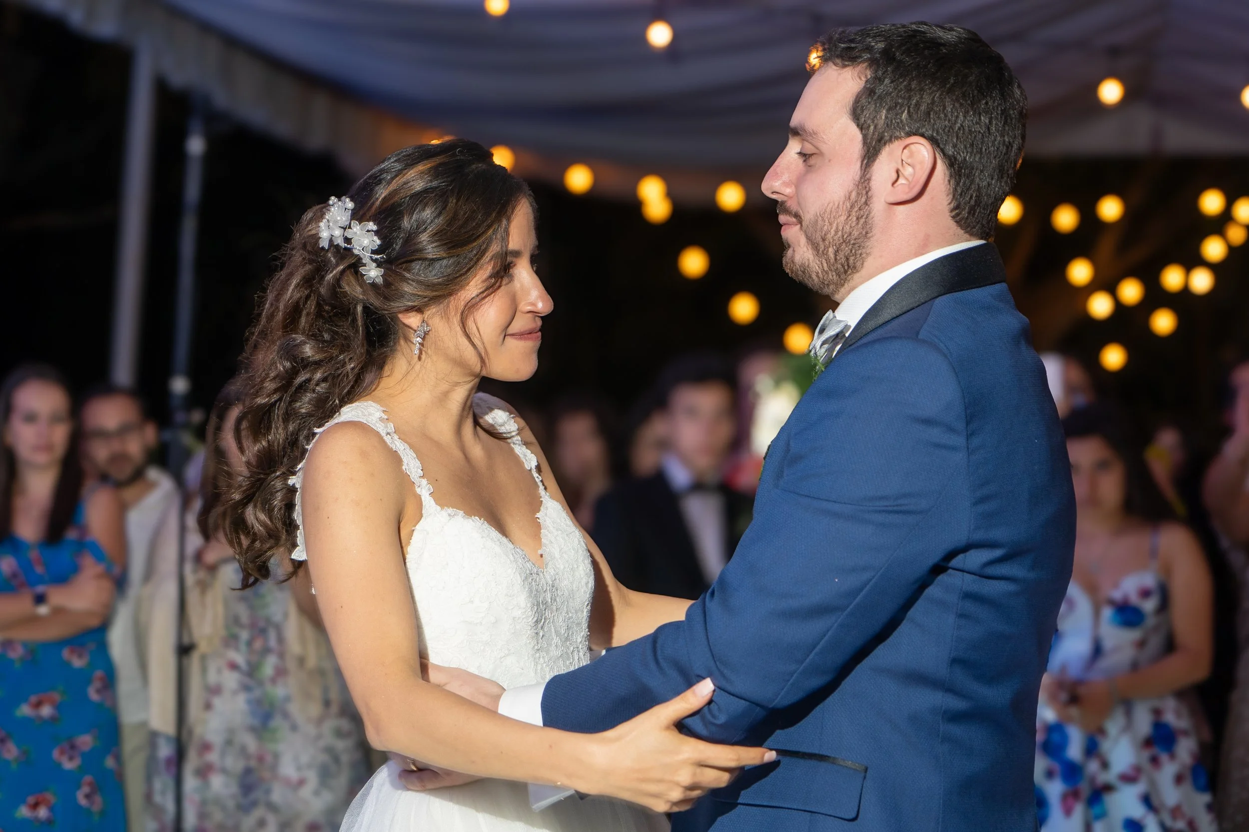 Pareja en la ceremonia de boda en un acto al aire libre, adultos en el fondo observando, decoraciones con luces cálidas.