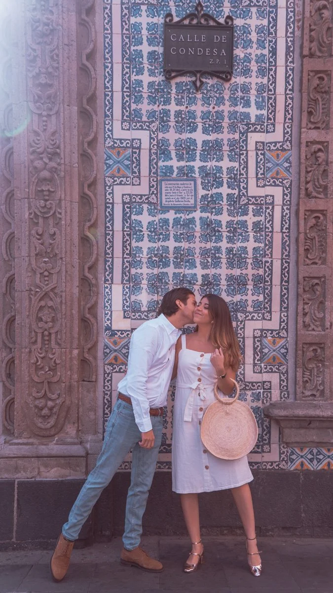A couple stands close, with the man kissing the woman's cheek in front of a decorated blue and white tiled wall with a street sign that says 'Calle de Condesa.' They are dressed in casual white outfits. The man wears jeans and brown shoes, and the wo