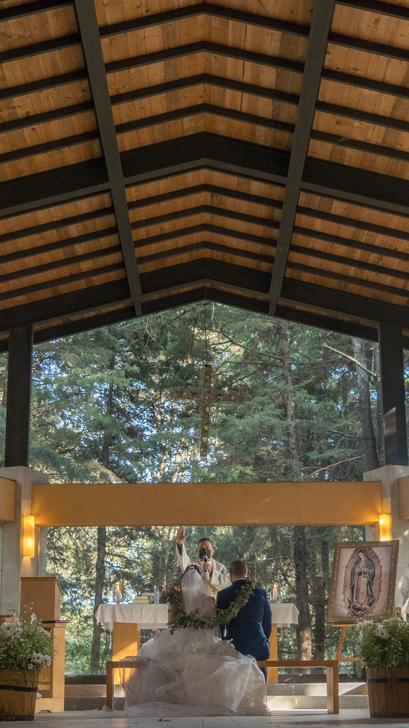 Durante una ceremonia en una iglesia, una pareja de novios está de espaldas, tocando el altar, con un sacerdote frente a ellos. La iglesia tiene un gran ventanal con un árbol y un crucifijo visible en el fondo. Hay flores decorativas y una imagen rel