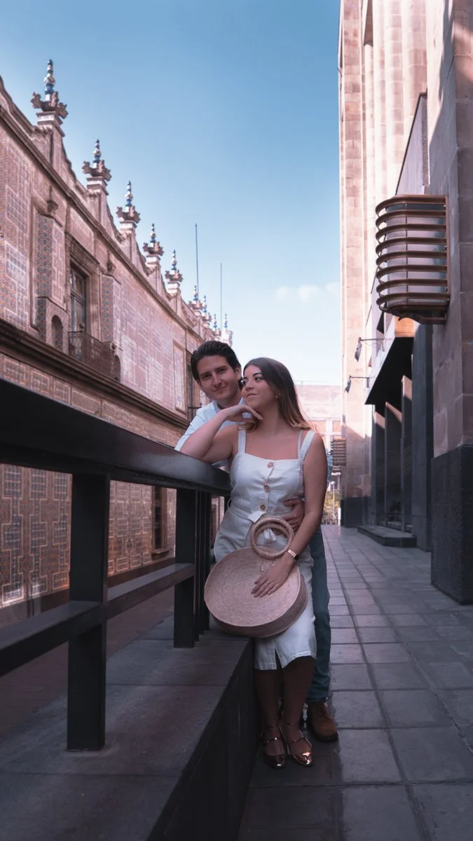 A young couple stands close together on a city street, with historic architecture and a blue sky in the background. The woman is holding a large straw hat and wearing a white dress, while the man stands behind her with his arm around her waist.