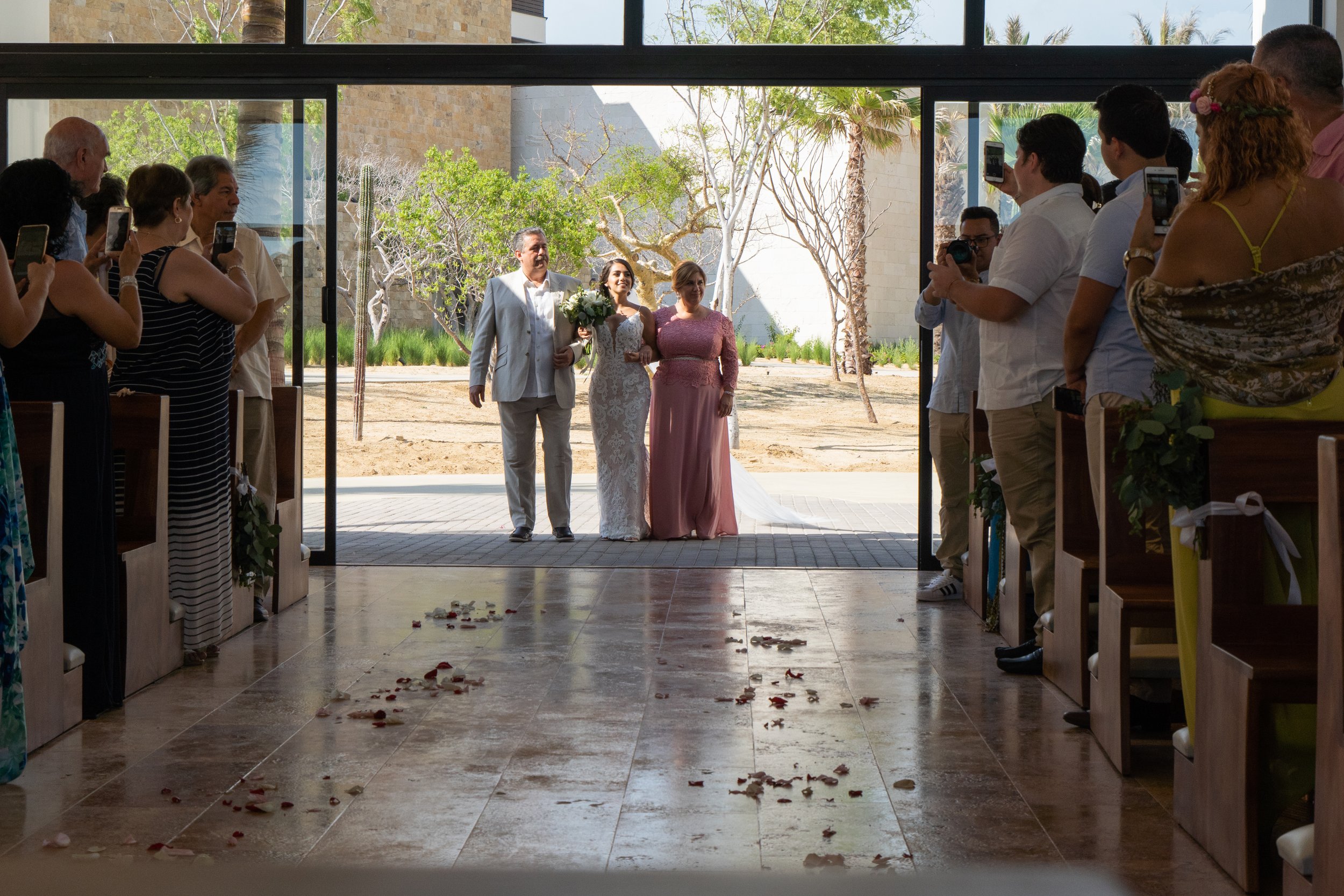 Boda en una iglesia con invitados tomando fotos, vista desde el interior hacia la entrada, con la pareja en el altar y decoraciones florales en las bancas.
