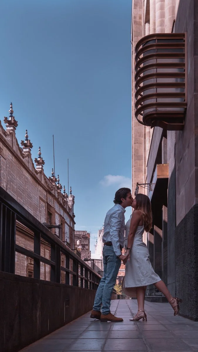 A couple kissing on a city sidewalk, holding hands, with historic building architecture and a clear blue sky in the background.