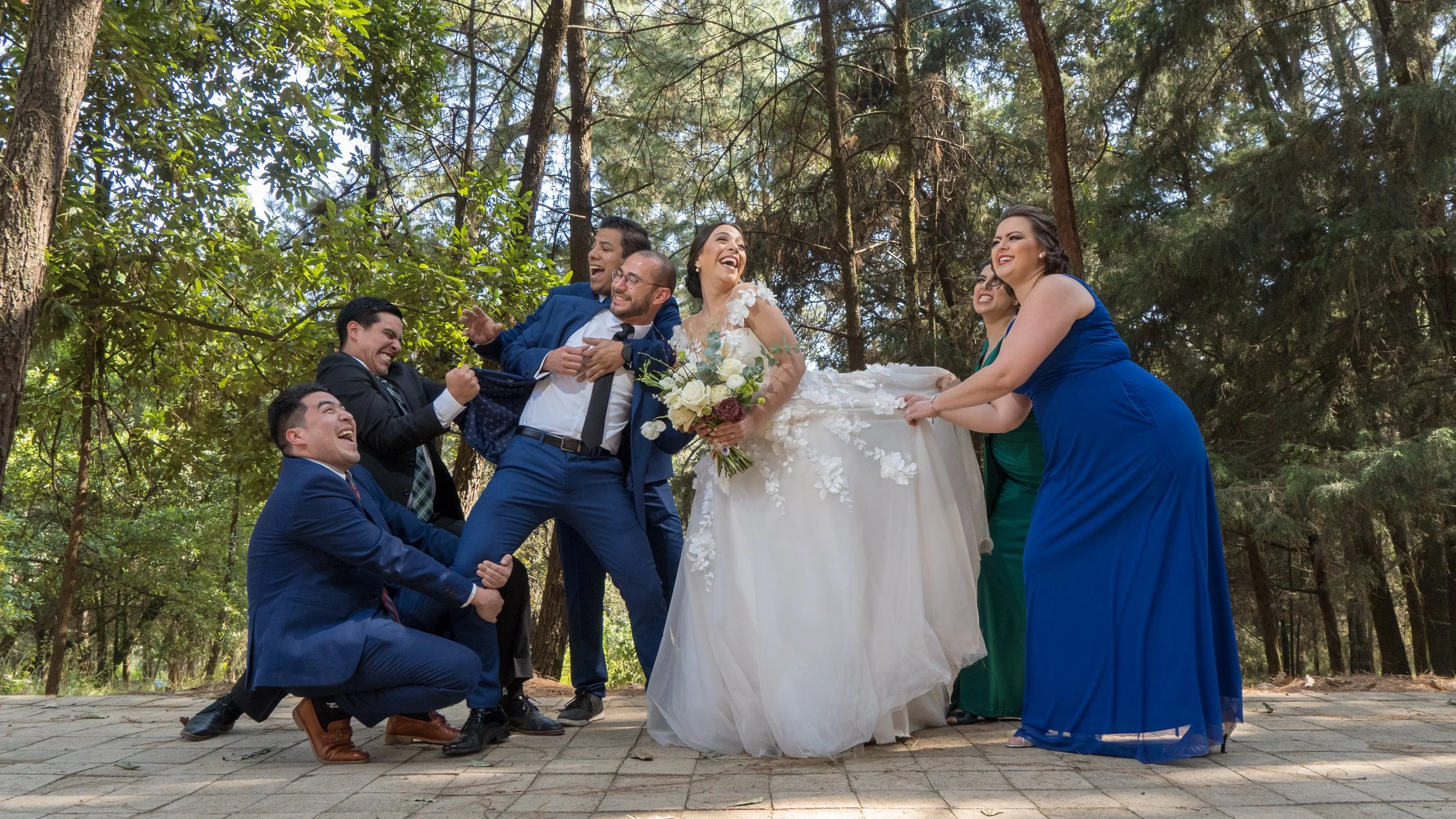 Grupo de siete personas en un bosque en una boda, riendo y divirtiéndose, con la novia en vestido blanco en el centro sosteniendo un ramo, rodeada de amigos en trajes y vestidos color azul y verde.
