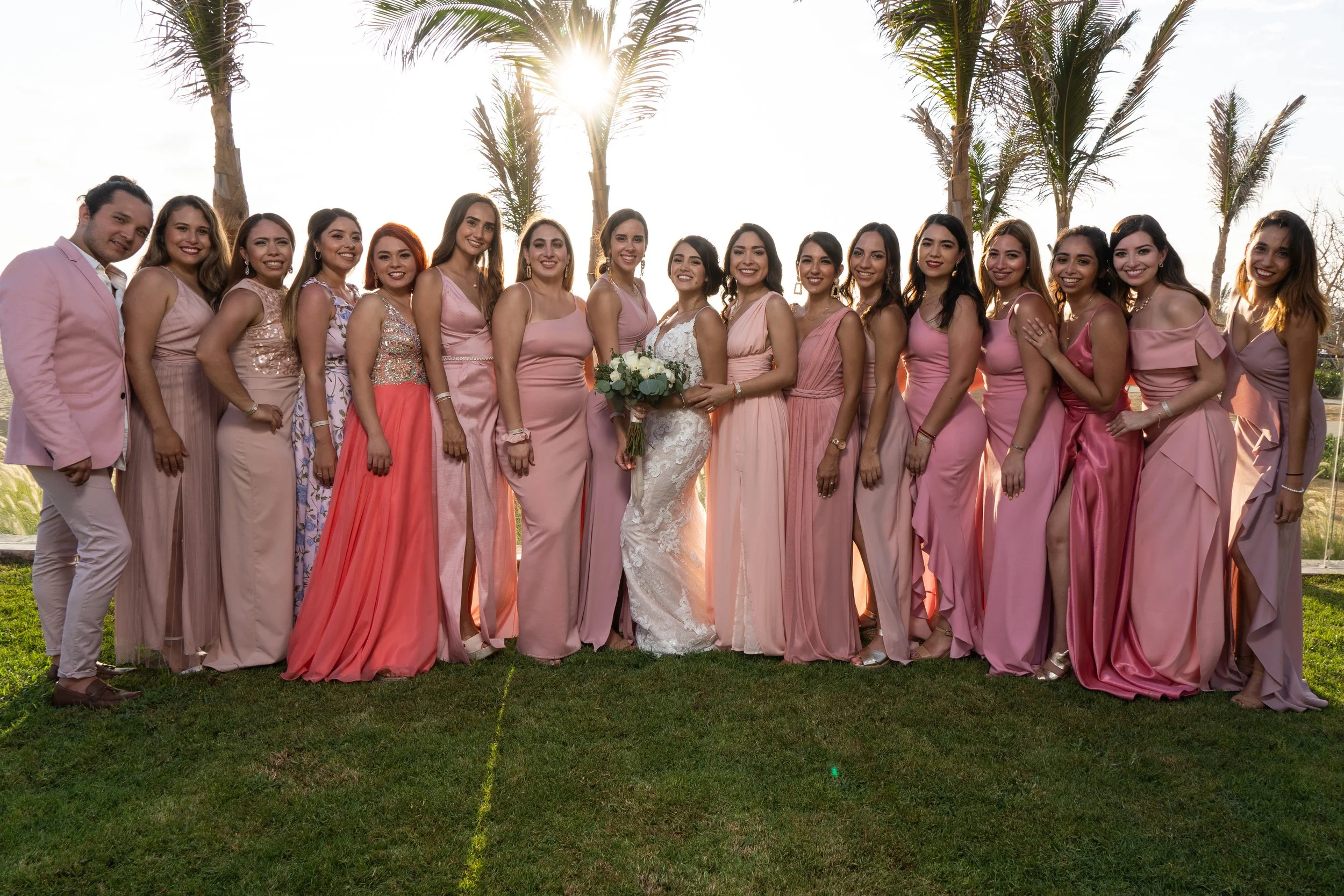 Grupo de personas en vestido formal y de colores rosados, con una mujer en vestido de novia en el centro, en un entorno al aire libre con palmeras, durante el atardecer.