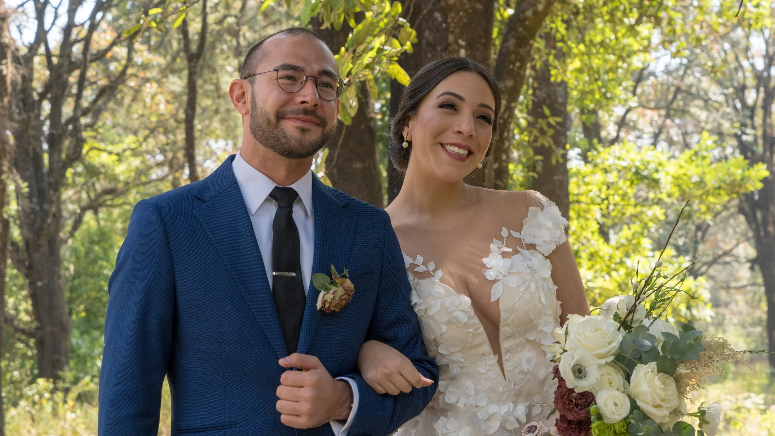 Pareja de recién casados caminando en un parque, la mujer sostiene un ramo de flores y ambos lucen felices con árboles y luz natural en el fondo.
