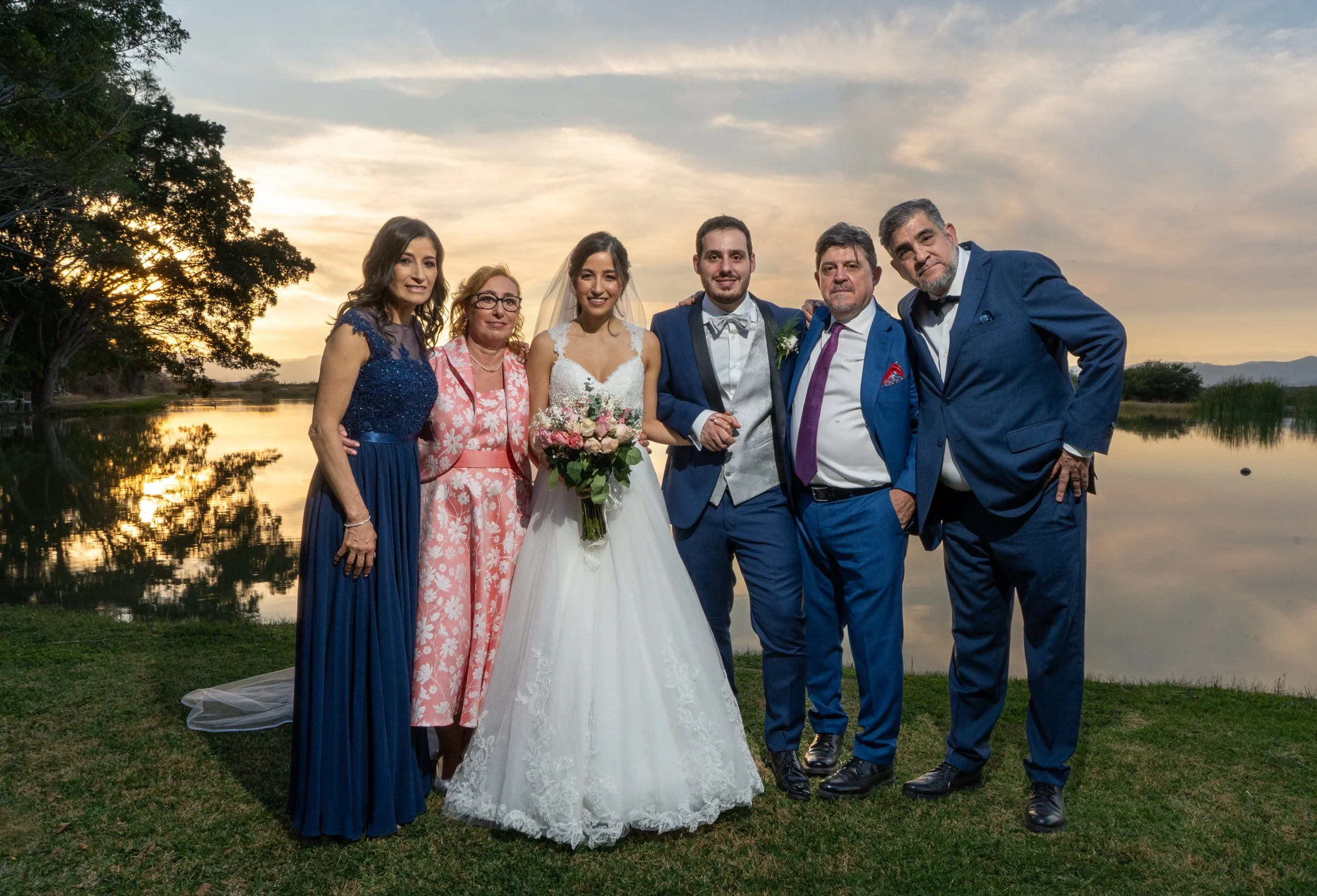 Grupo de personas en una boda al atardecer junto a un lago con árboles y montañas en el fondo.