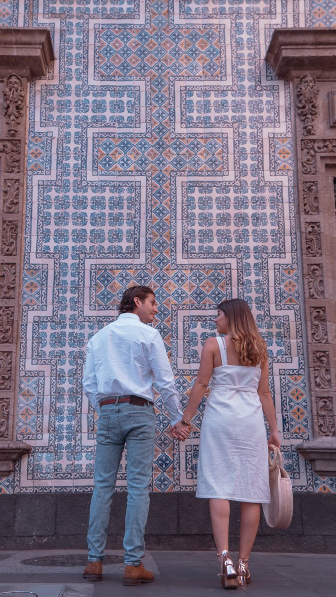 A couple holding hands and looking at each other in front of a decorative tiled wall, with ornate columns on either side.