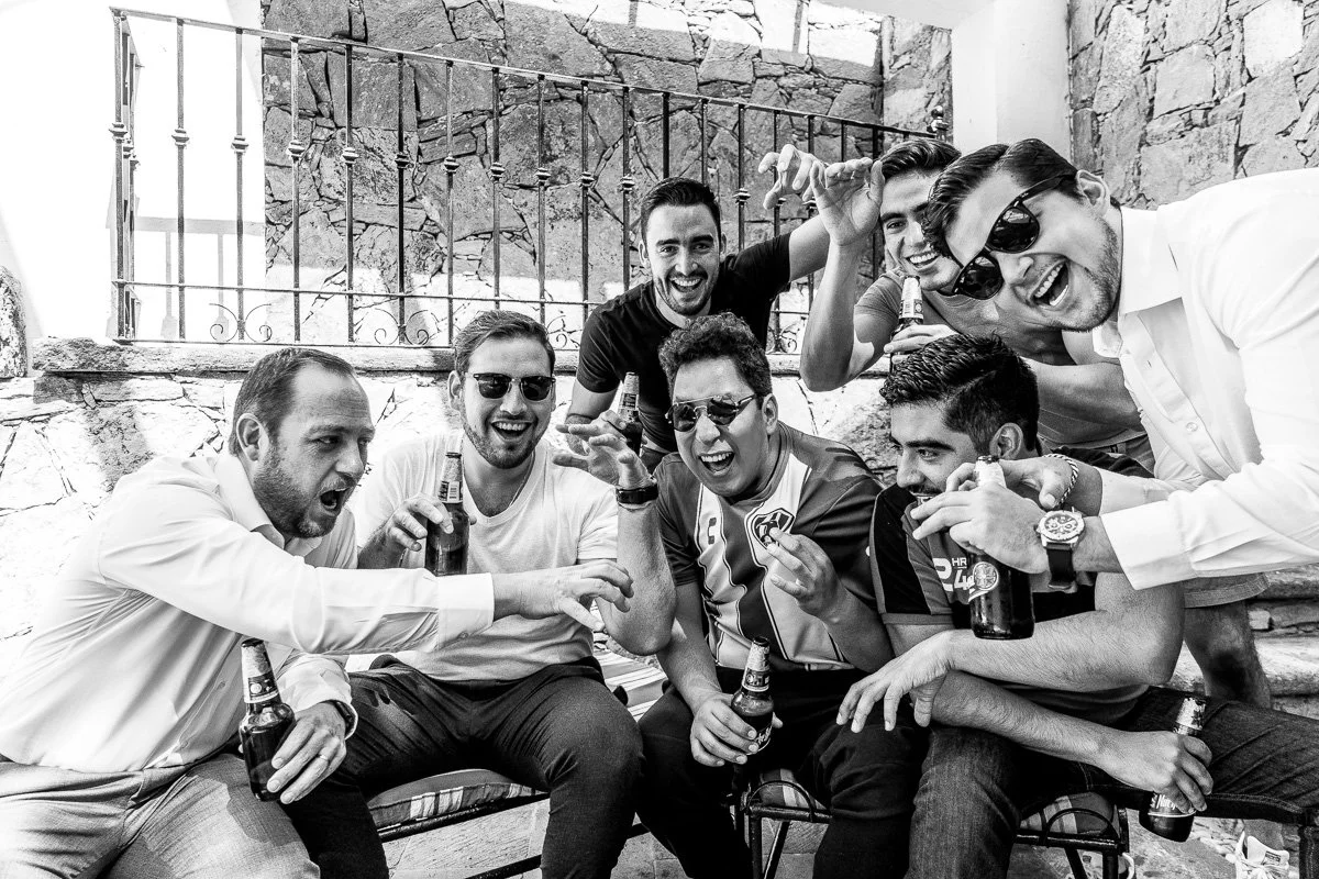 A group of men celebrating, holding beer bottles, smiling, some wearing sunglasses, in front of a stone wall with a metal fence.