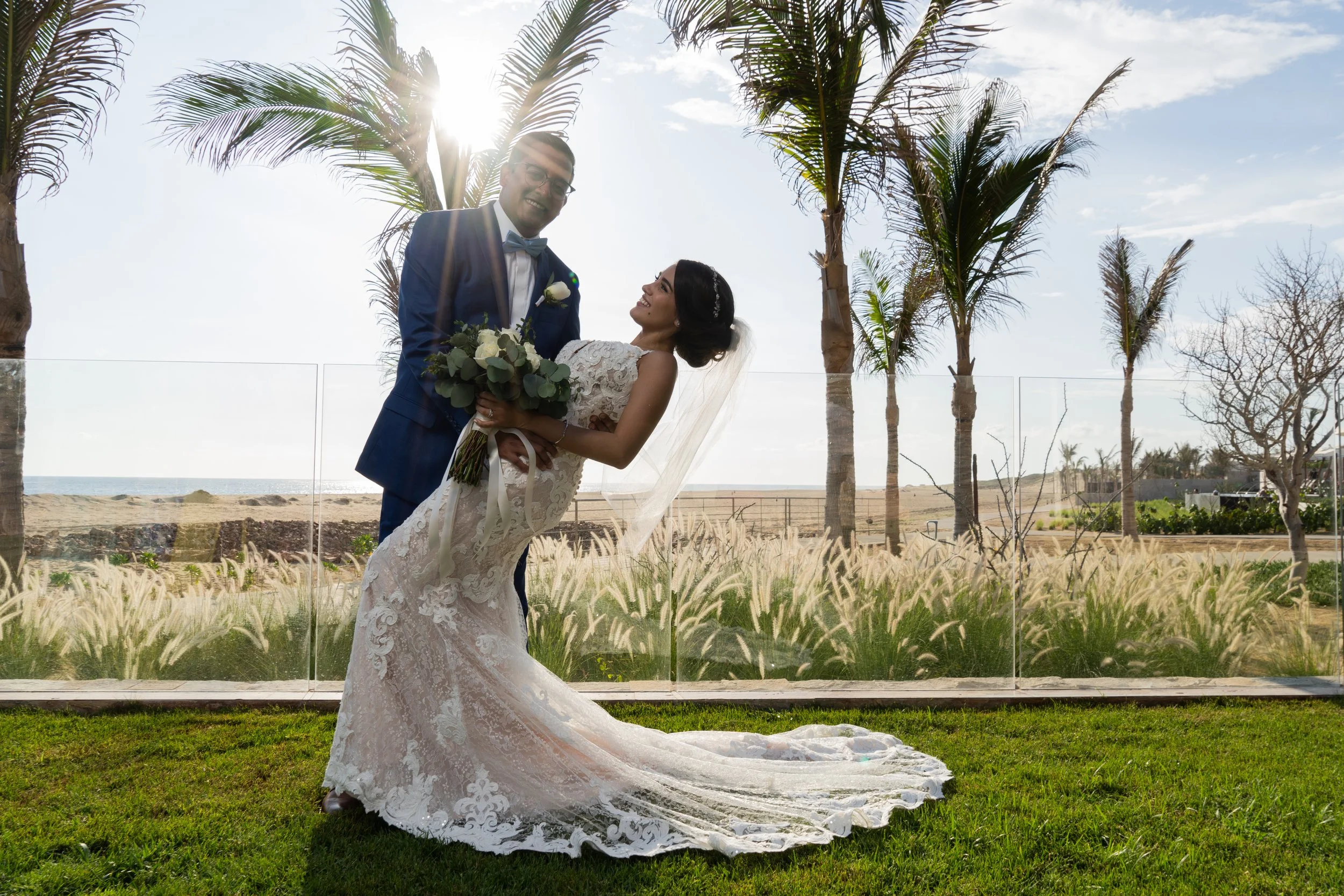 Pareja de recién casados en un jardín con palmeras, ella con vestido de novia y él con traje azul, durante una sesión fotográfica al atardecer.