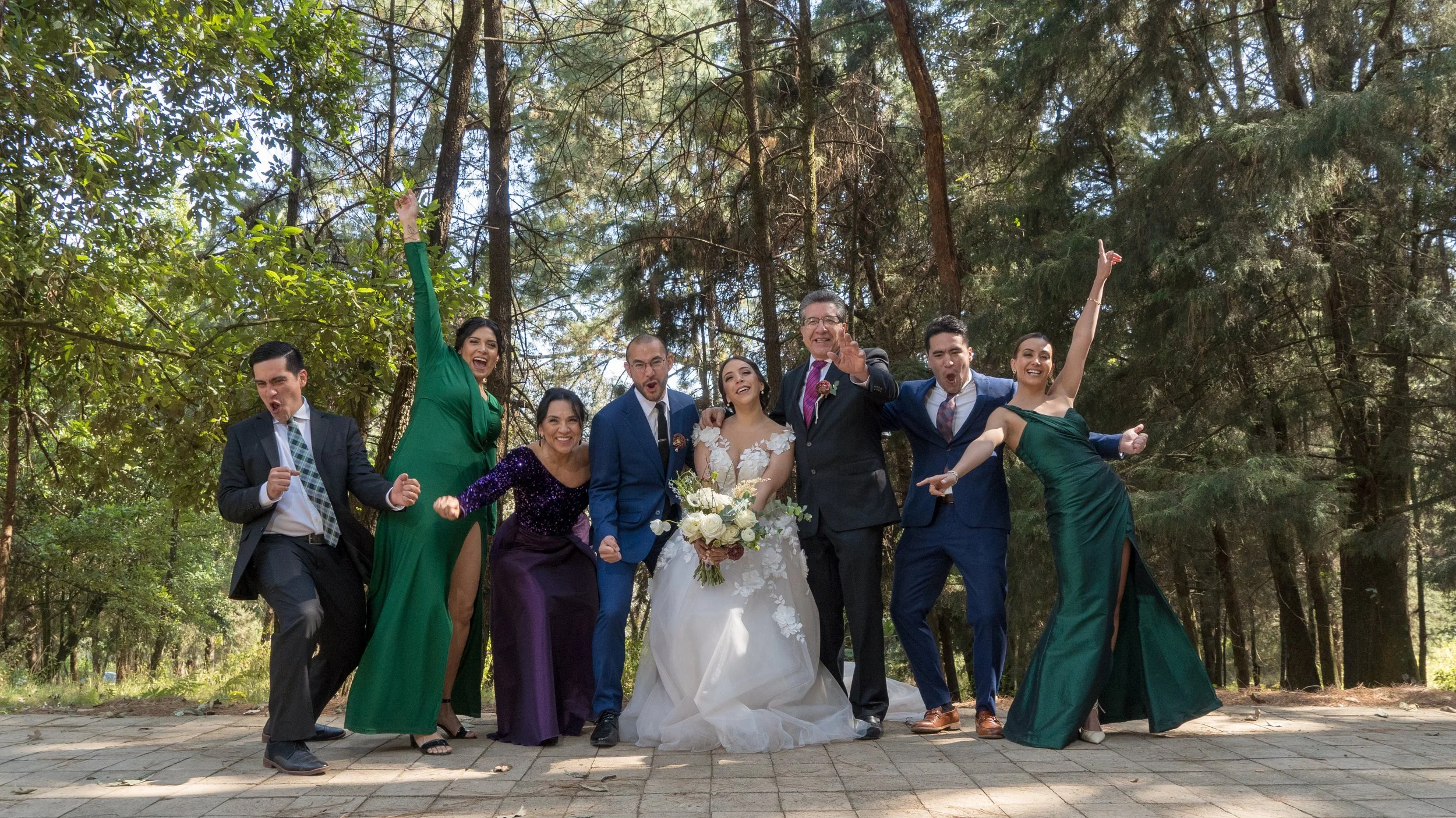 Grupo de diez personas en celebración, posando alegremente en un entorno natural con árboles de fondo, en un evento formal o boda, algunos con trajes y vestidos elegantes, todos con expresiones de felicidad y entusiasmo.