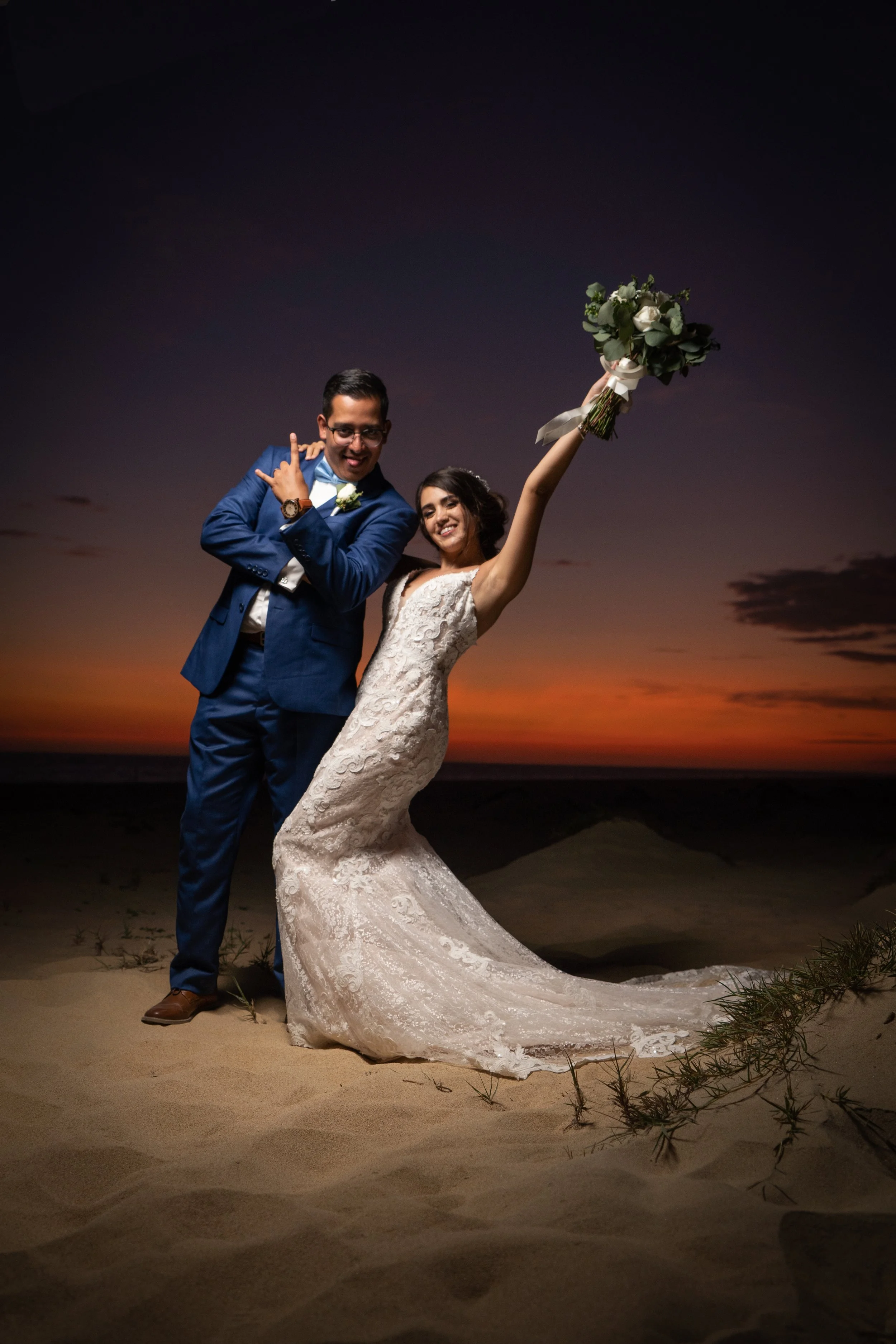 Pareja de novios en la playa al atardecer, la novia sostiene un ramo y sonríe, el novio hace una pose divertida junto a ella.