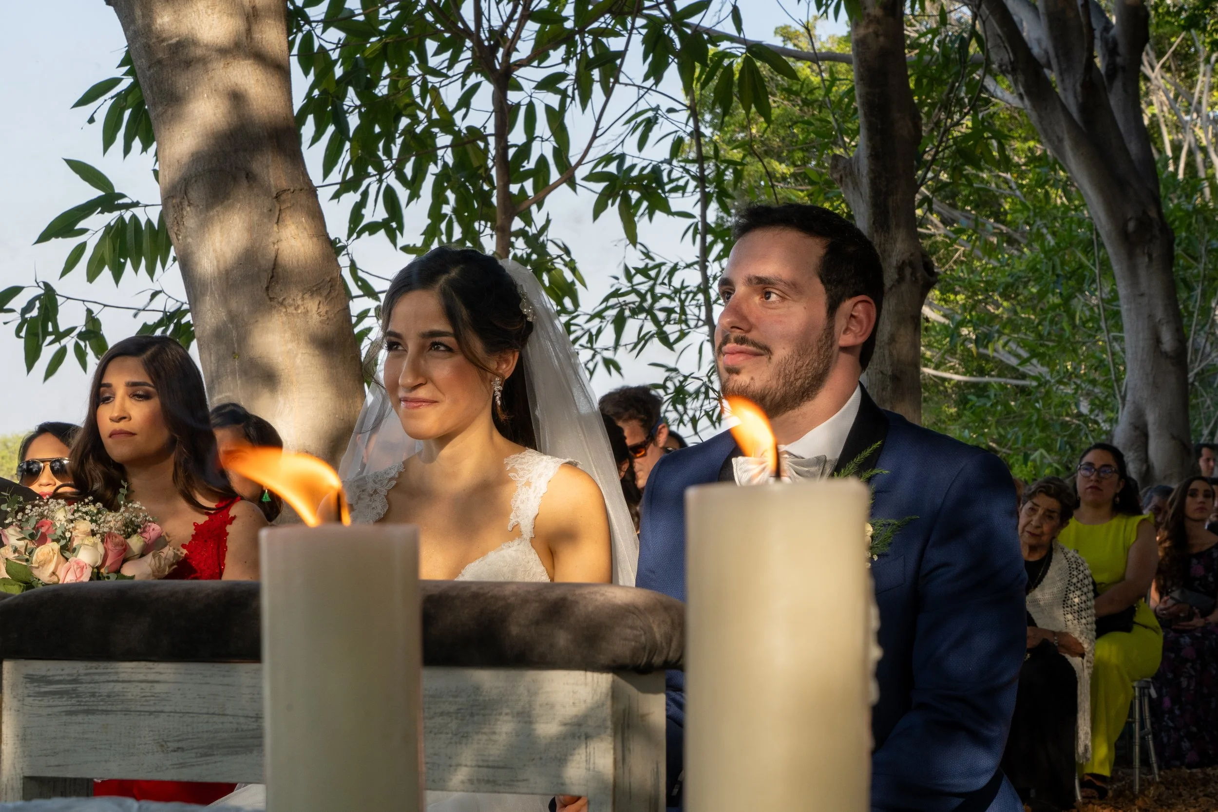 Pareja de novios en una boda al aire libre, rodeados de invitados, con árboles y velas en primer plano.