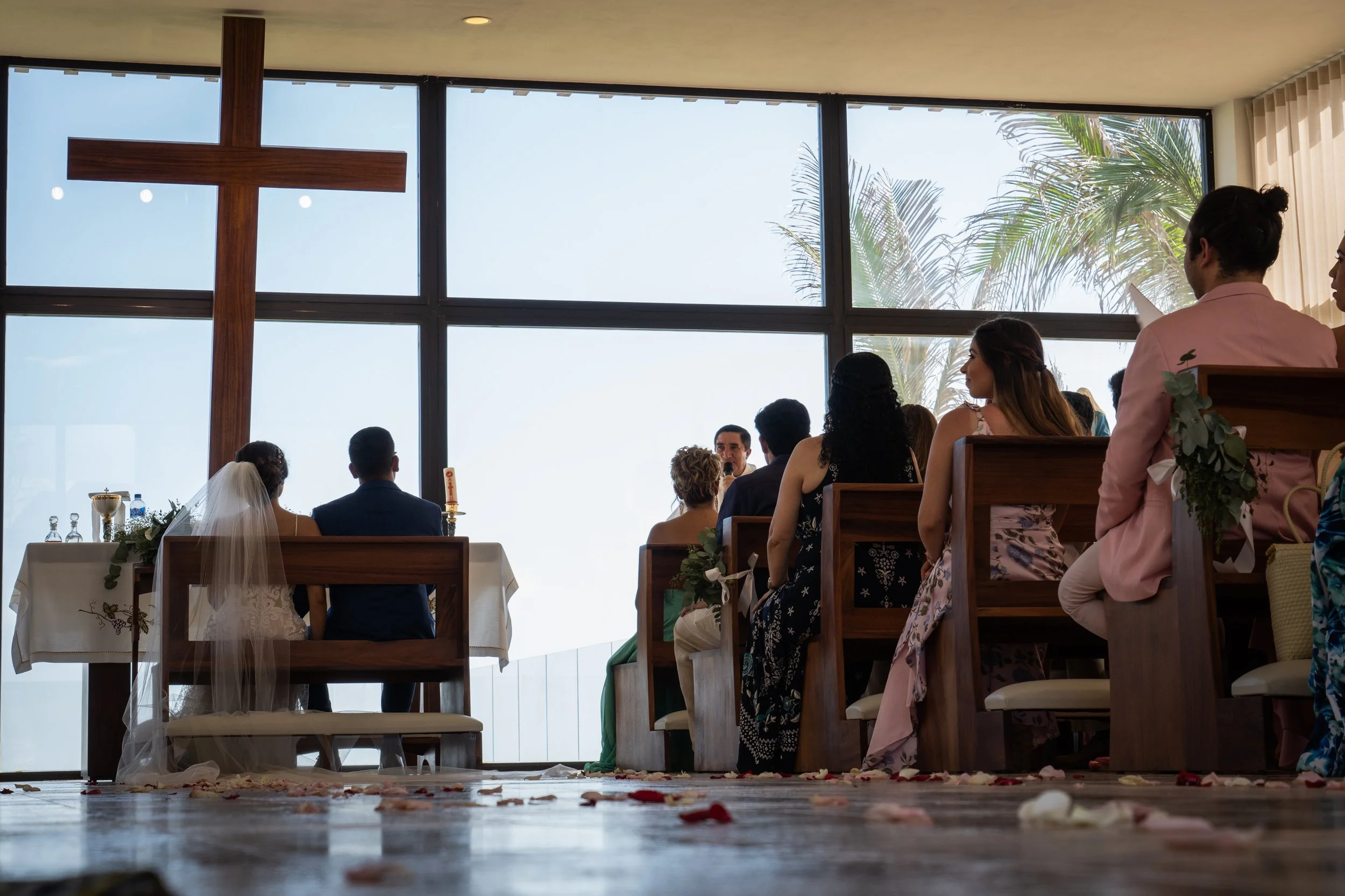 Ceremonia religiosa en una iglesia con cruz de madera y gran ventana con vista al cielo y palmeras, donde una pareja se casa con invitados observando.
