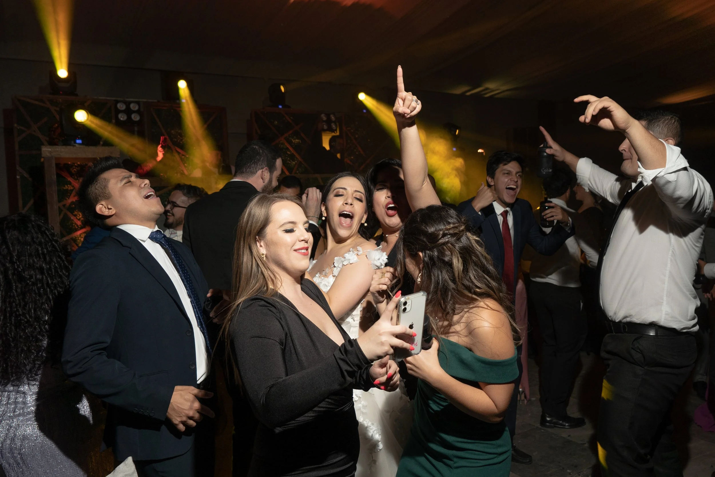 Personas en una fiesta bailando, cantando y tomando fotos, con luces y ambiente festivo.