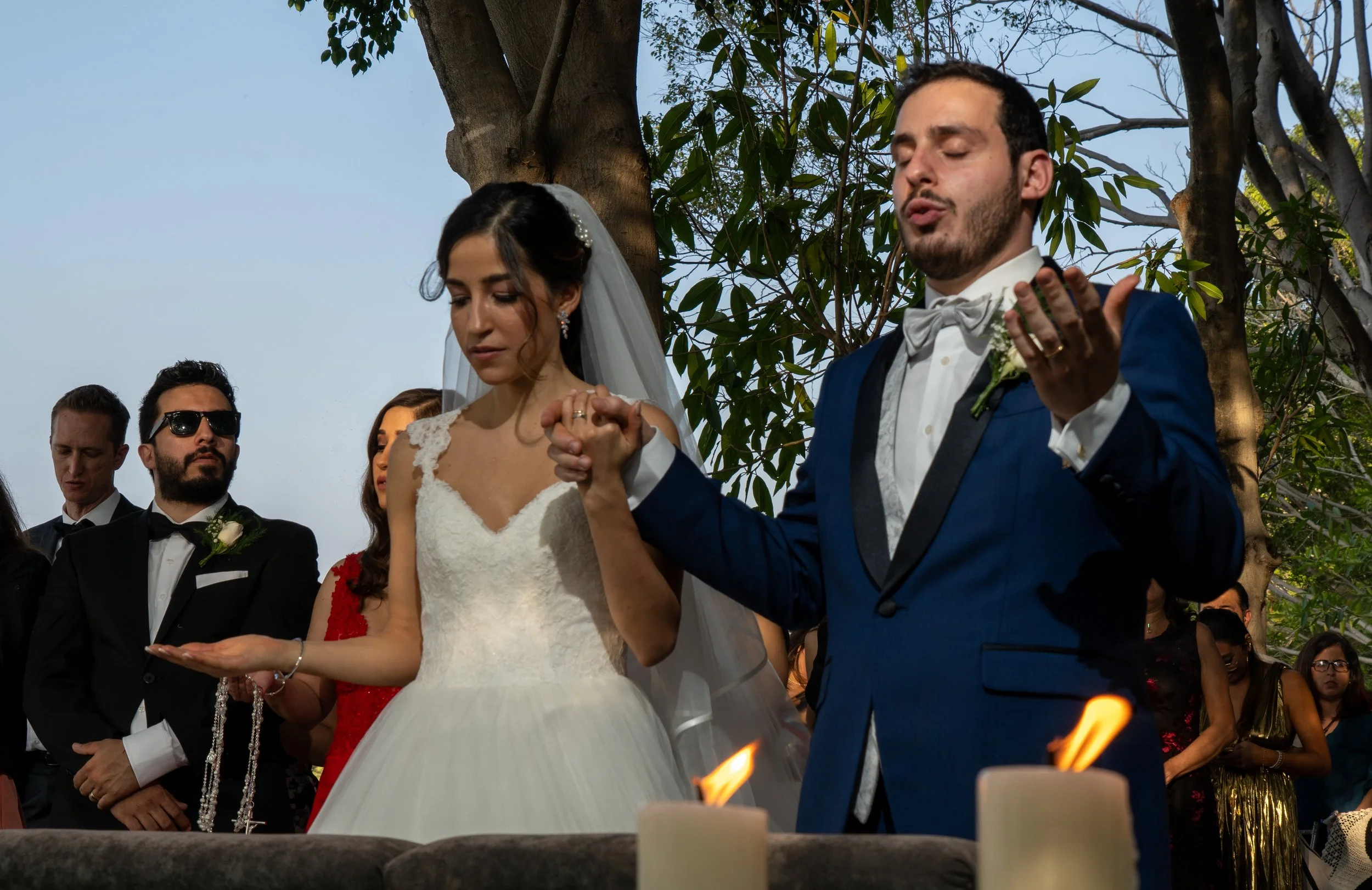 Boda al aire libre con la pareja principal rezando, rodeados de invitados, con árboles y cielo en el fondo.