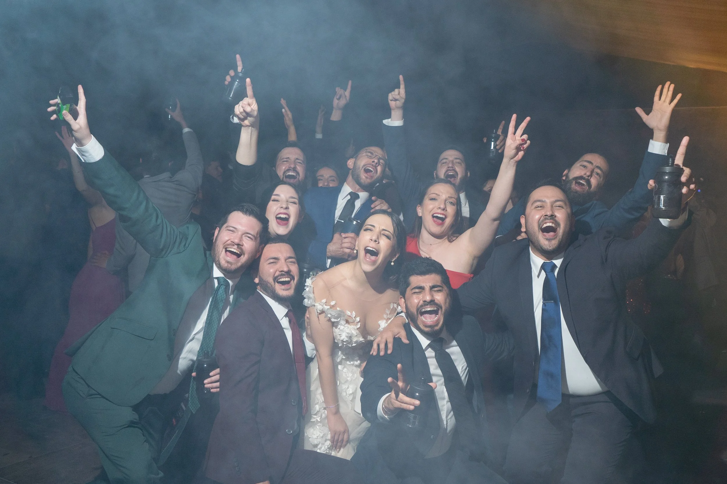 Grupo de personas celebrando en una fiesta, riendo y levantando las manos, algunas sosteniendo bebidas, ambiente alegre y festivo.