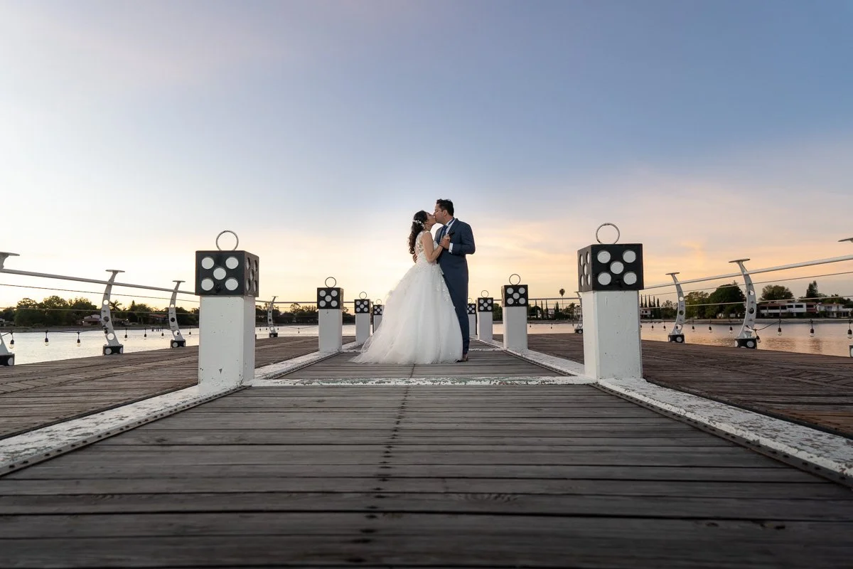 Couple in wedding attire standing on a wooden dock at sunset, facing each other and holding hands.