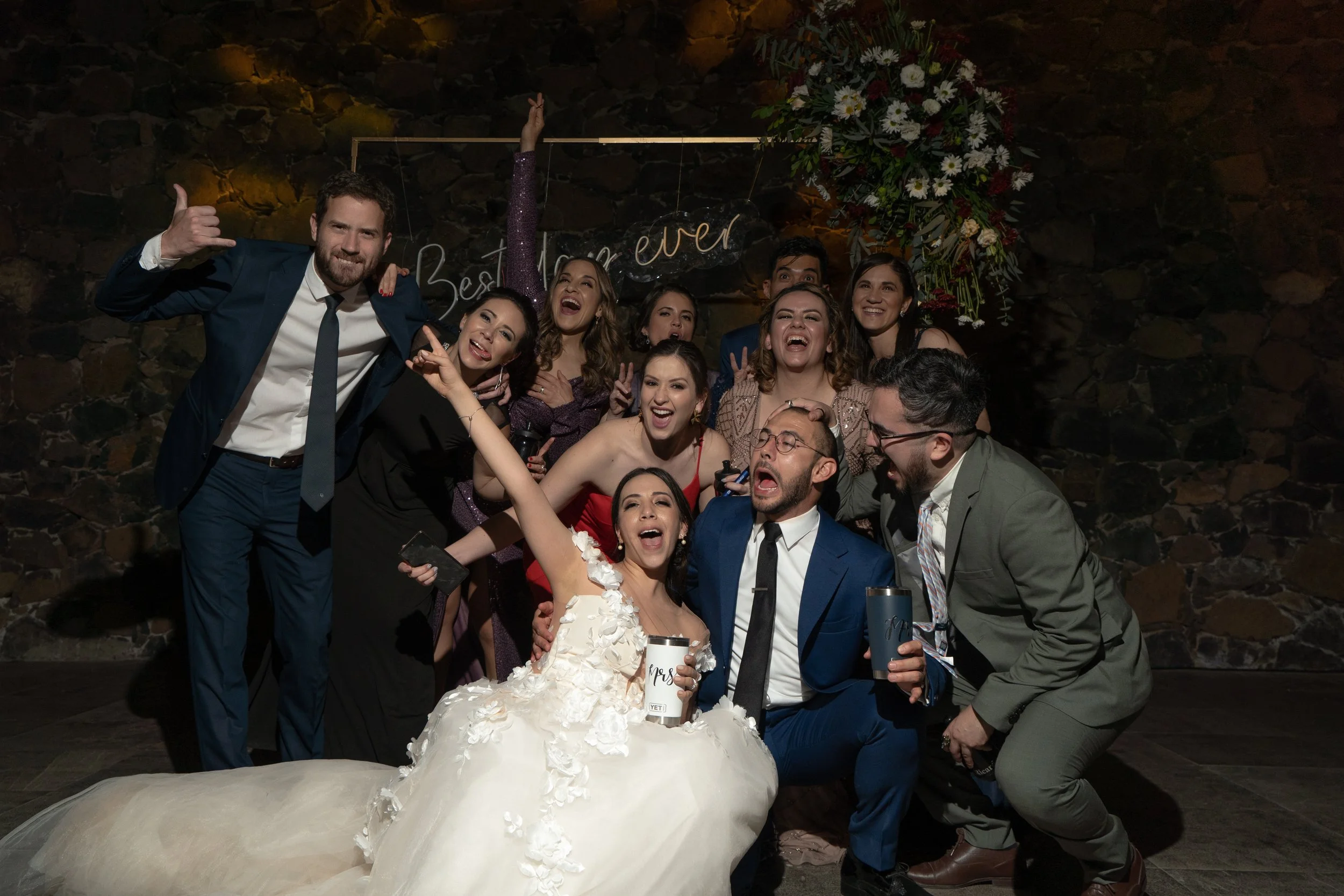 Grupo de personas en una boda, sonriendo y posando para la foto, con la novia en el centro con vestido blanco y flor en el cabello, rodeada de amigos y familiares vestidos formalmente.
