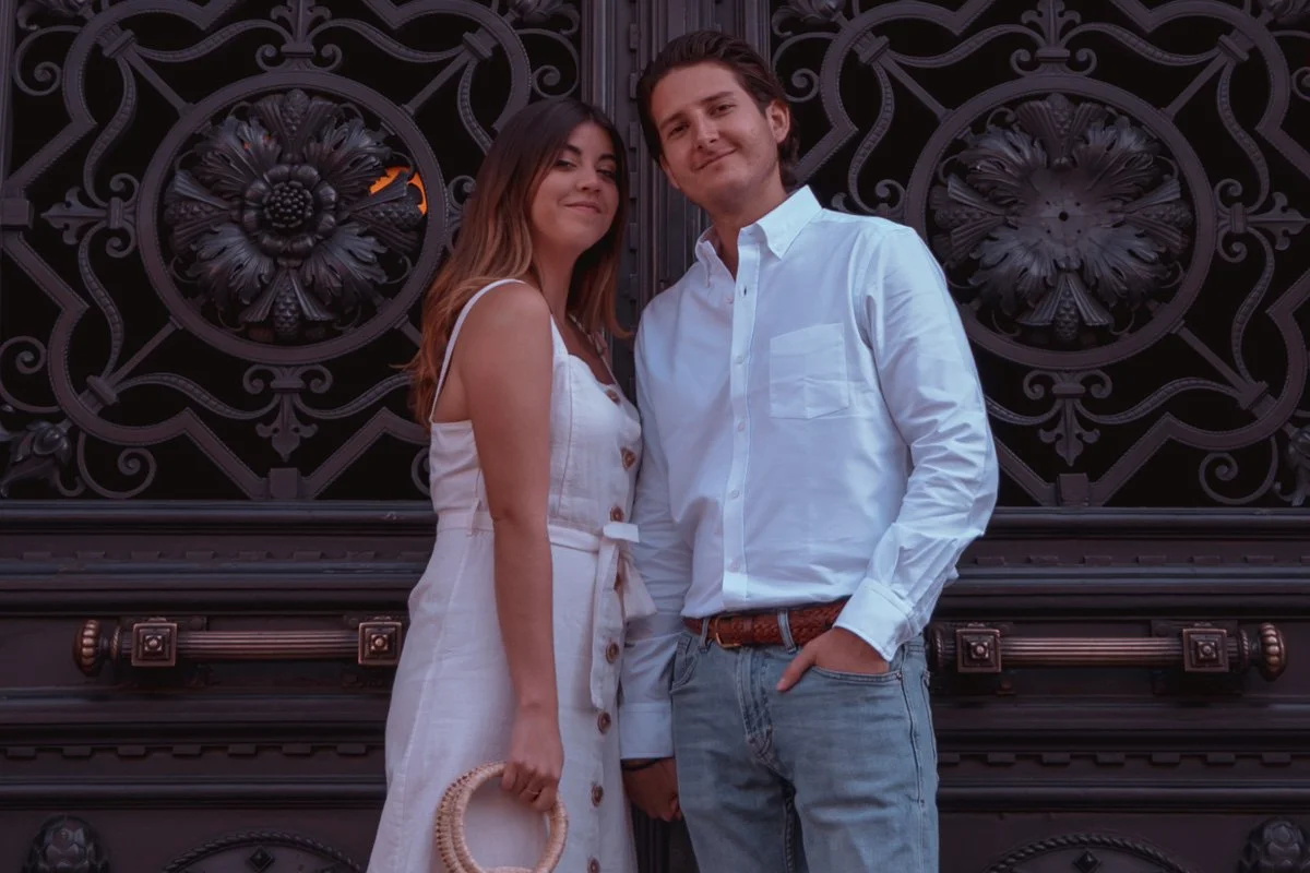 A young woman and man stand side by side in front of an iron gate with floral designs, posing for the photo. The woman is wearing a white dress with buttons and has long hair, holding a woven handbag. The man is wearing a white button-up shirt and li