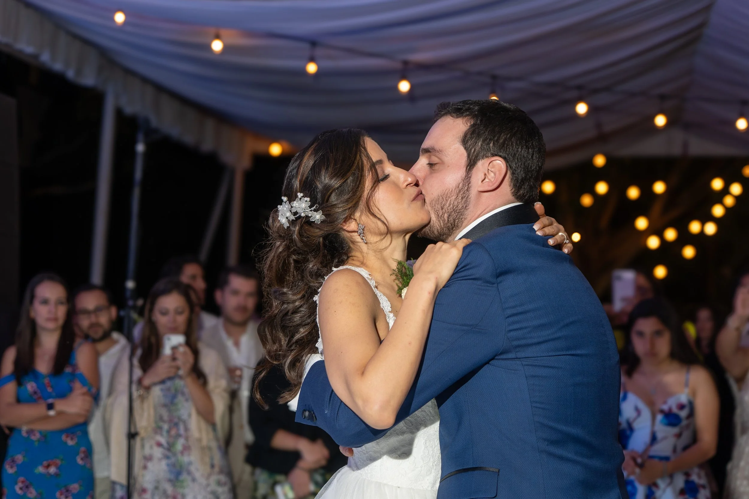 Pareja baila en una boda, rodeados de invitados y luces decorativas en el fondo.