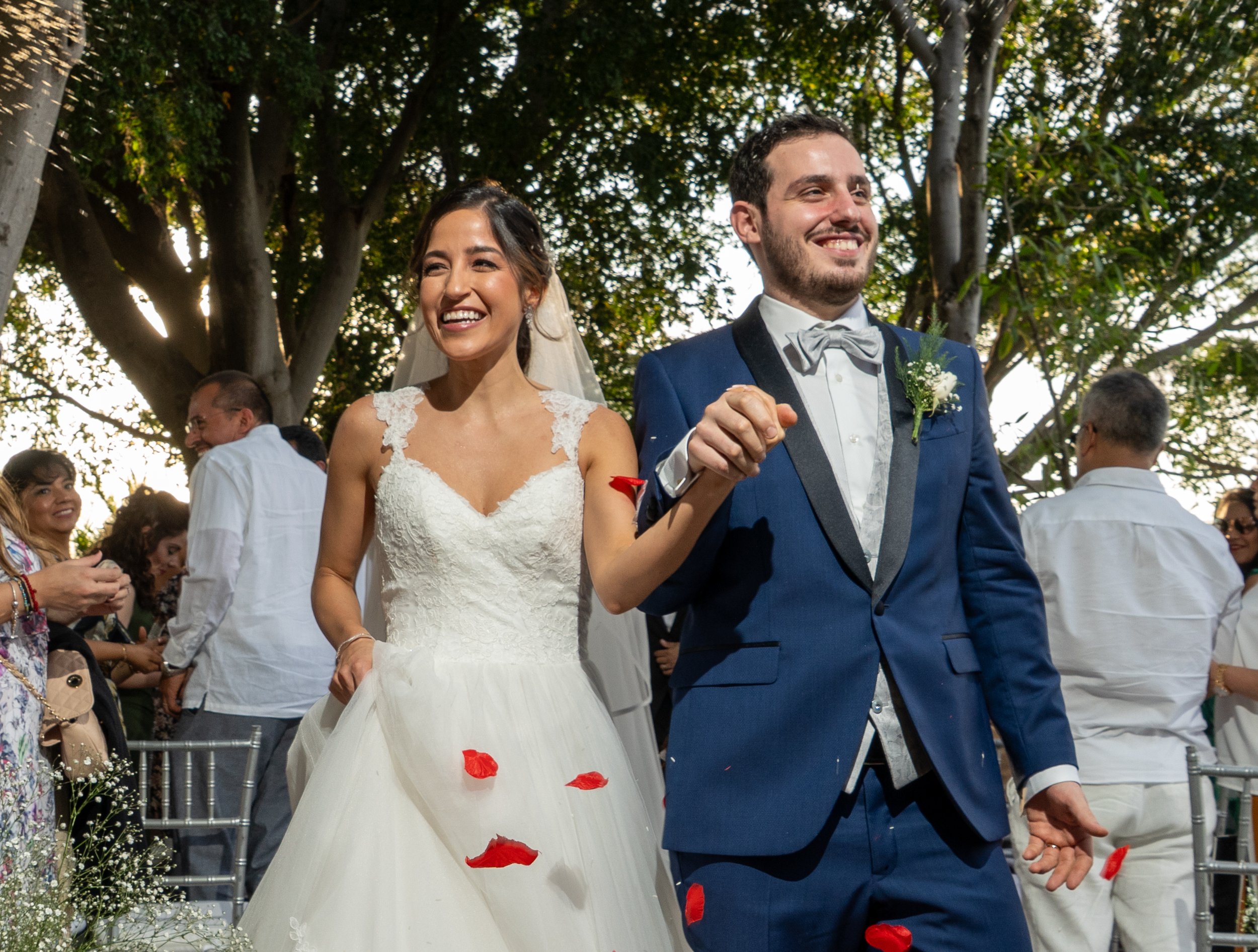 Pareja de novios en su boda, caminando juntos bajo árboles con invitados a su alrededor, mientras lanzan pétalos de rosa.