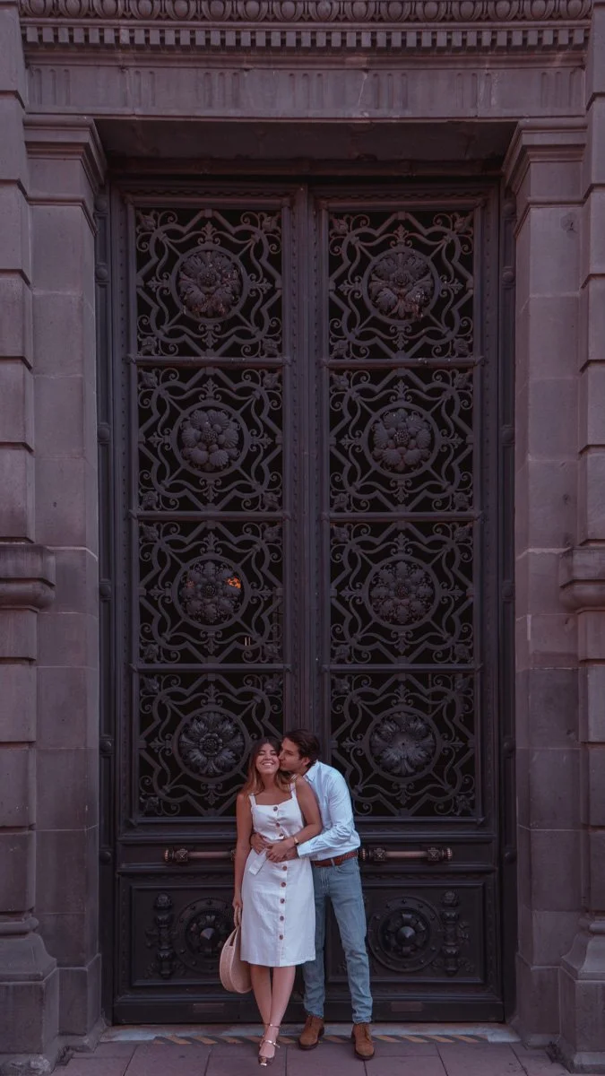 A couple standing in front of an ornate, large black iron gate with floral designs, part of a stone building facade.