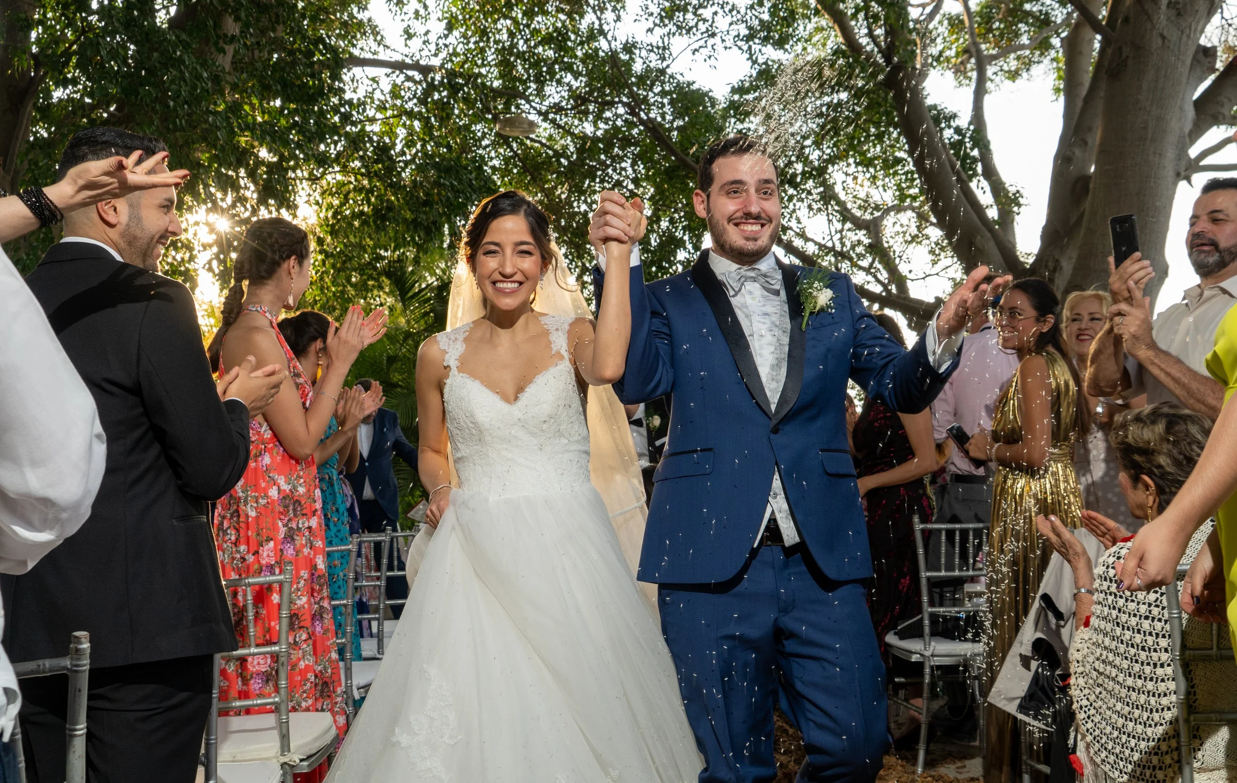 Pareja de novios en su boda, el hombre en traje azul y la mujer en vestido blanco, celebrando entre invitados en un entorno al aire libre con árboles, con confeti en el aire.