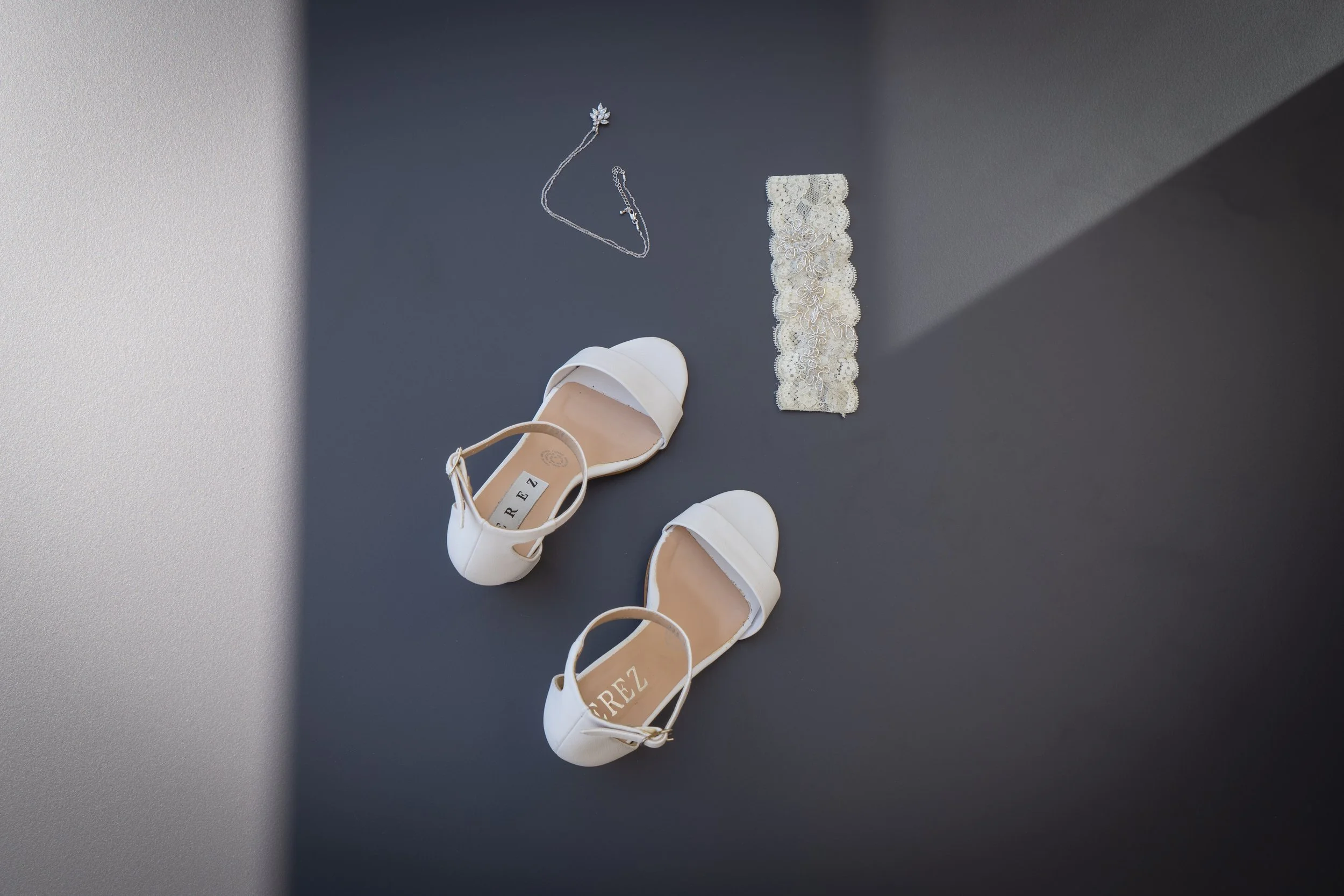 Zapatos blancos de tacón bajo, una cadena de plata, y un lazo de encaje en una superficie de color gris oscuro con luz natural.