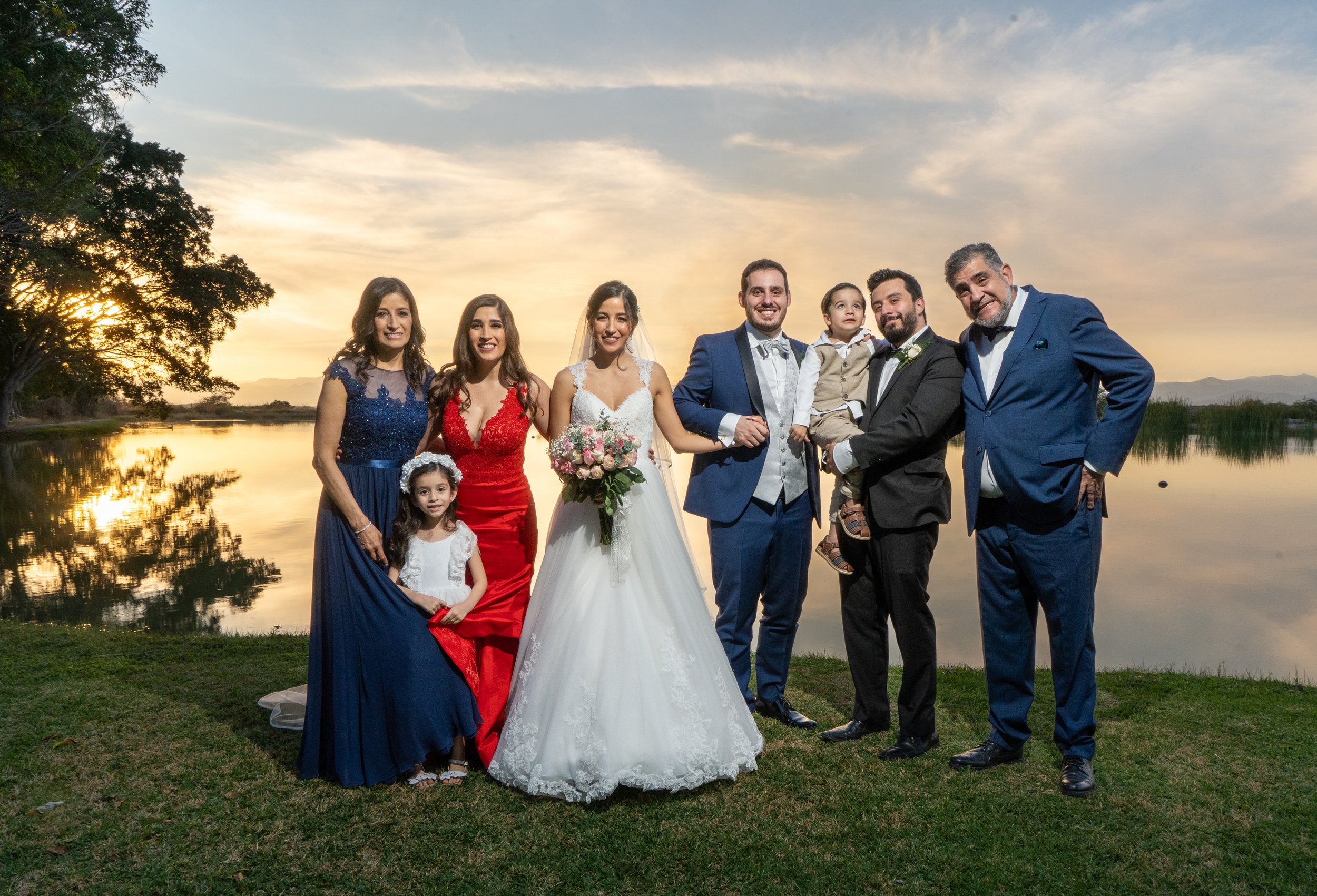 Grupo de personas en boda junto a un río al atardecer, con vestidos elegantes y sonriendo.