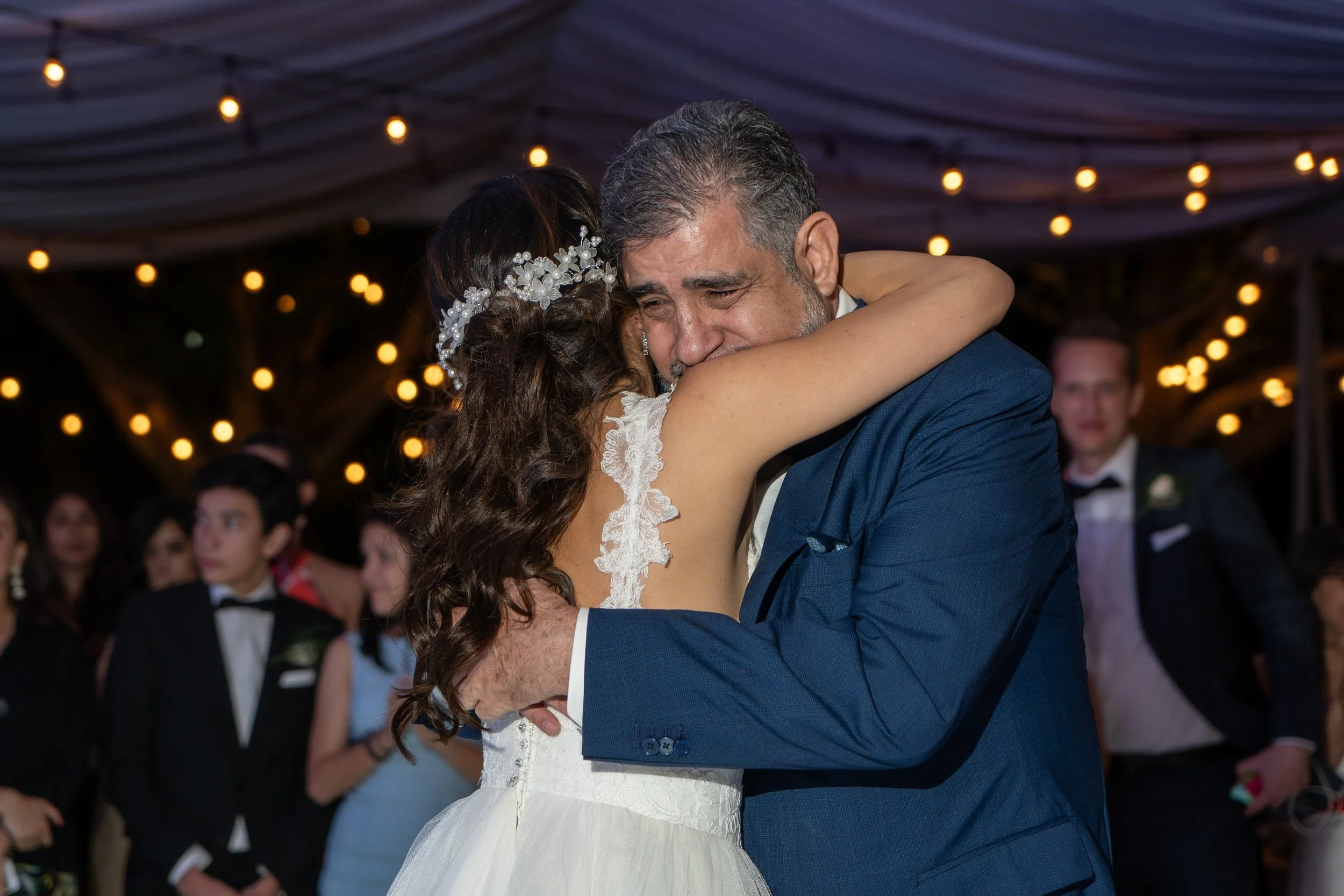 Una mujer y un hombre mayores, vestidos de formal, se están abrazando emocionados en una boda, con personas en el fondo y luces decorativas colgando encima.