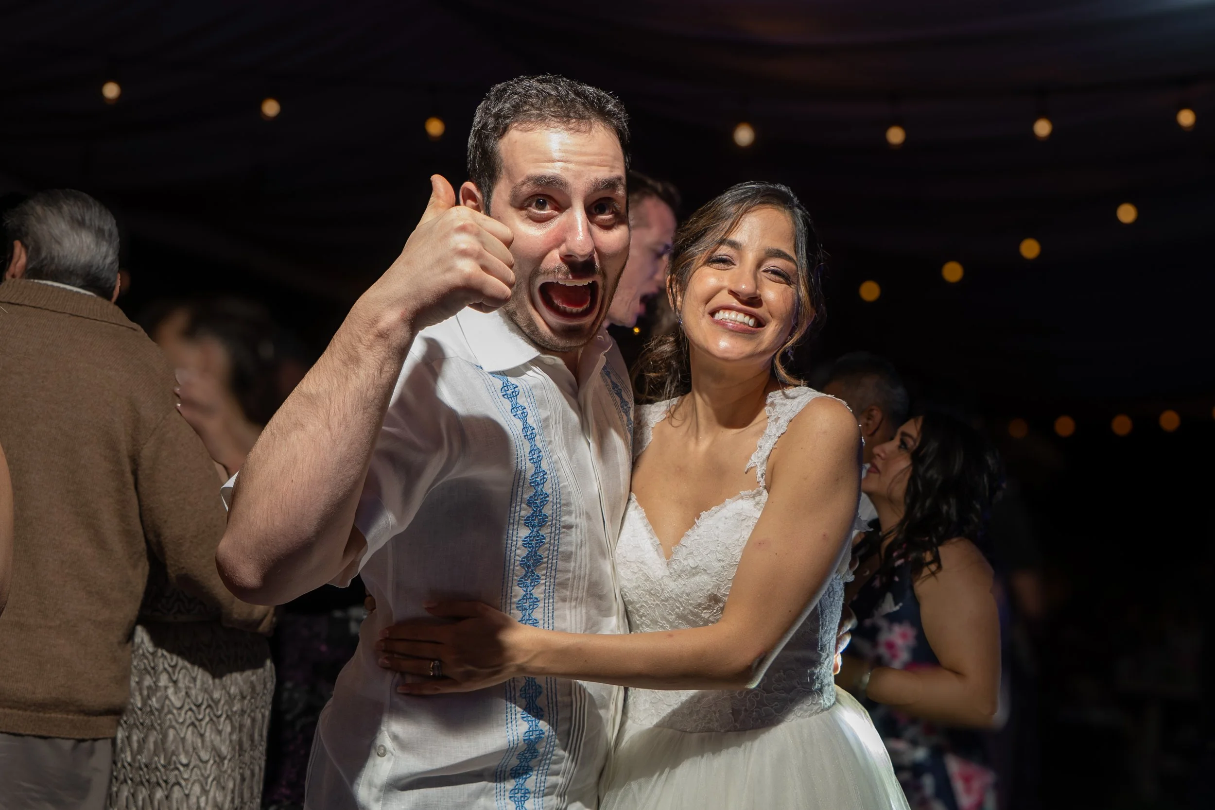 Pareja de recién casados bailando y sonriendo durante una celebración en un entorno oscuro con luces decorativas