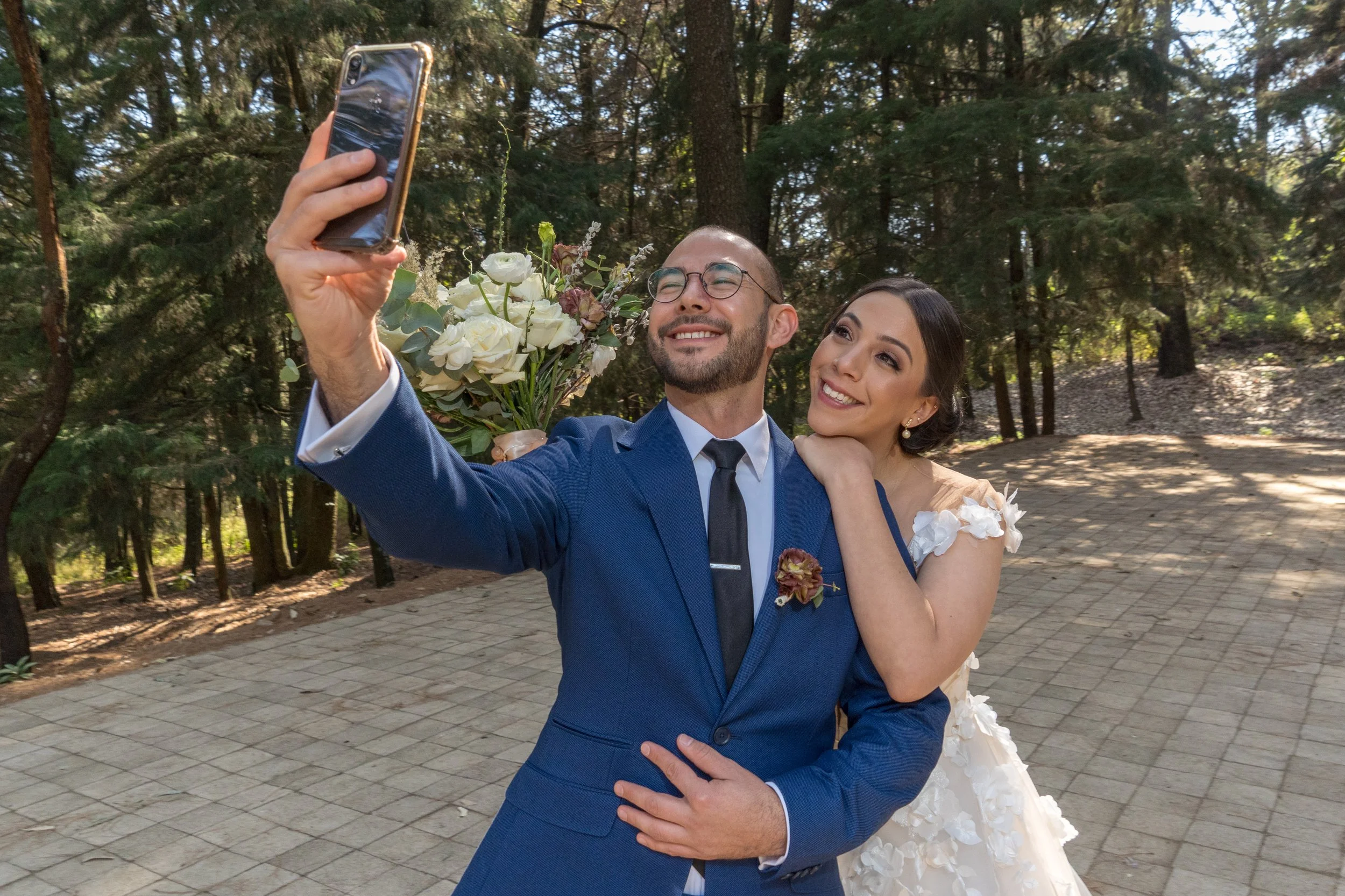 Una pareja de recién casados tomándose una selfie en un parque con árboles y suelo de piedra, el hombre sostiene un ramo de flores y ambos sonríen.