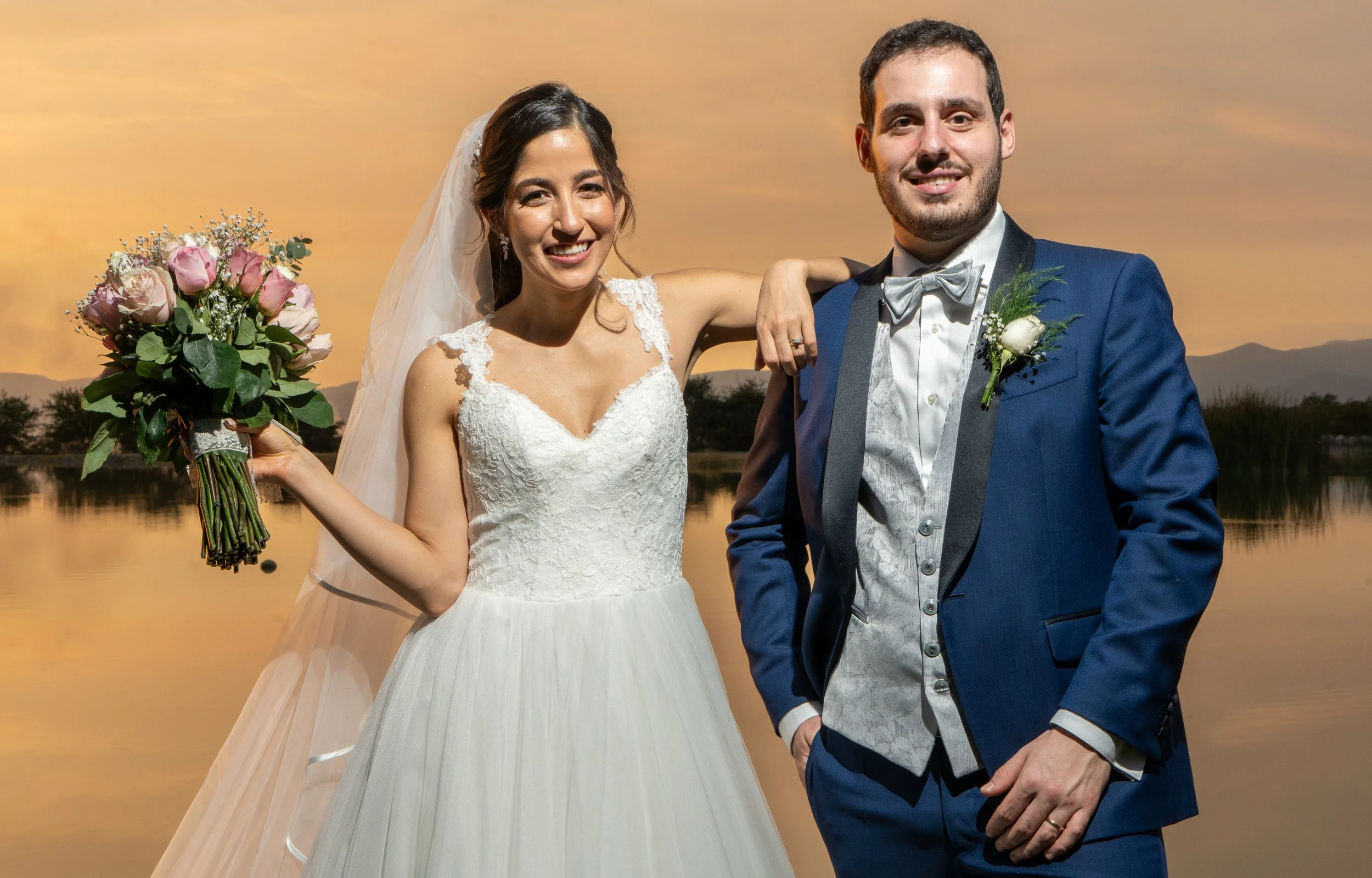 una pareja de recién casados en un atardecer, la mujer con vestido blanco y florero y el hombre con traje azul y pajarita, ambos sonriendo