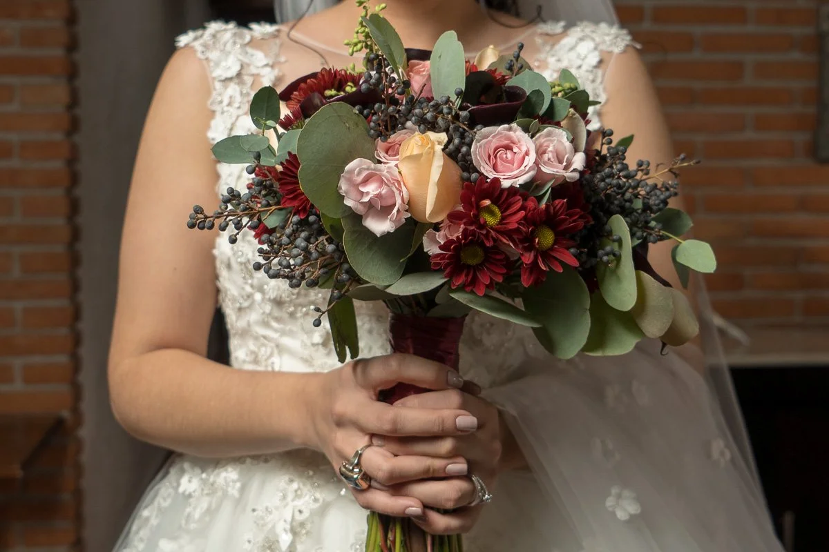 A bride holding a bouquet of flowers, including roses, dahlias, and greenery, in front of a brick wall.