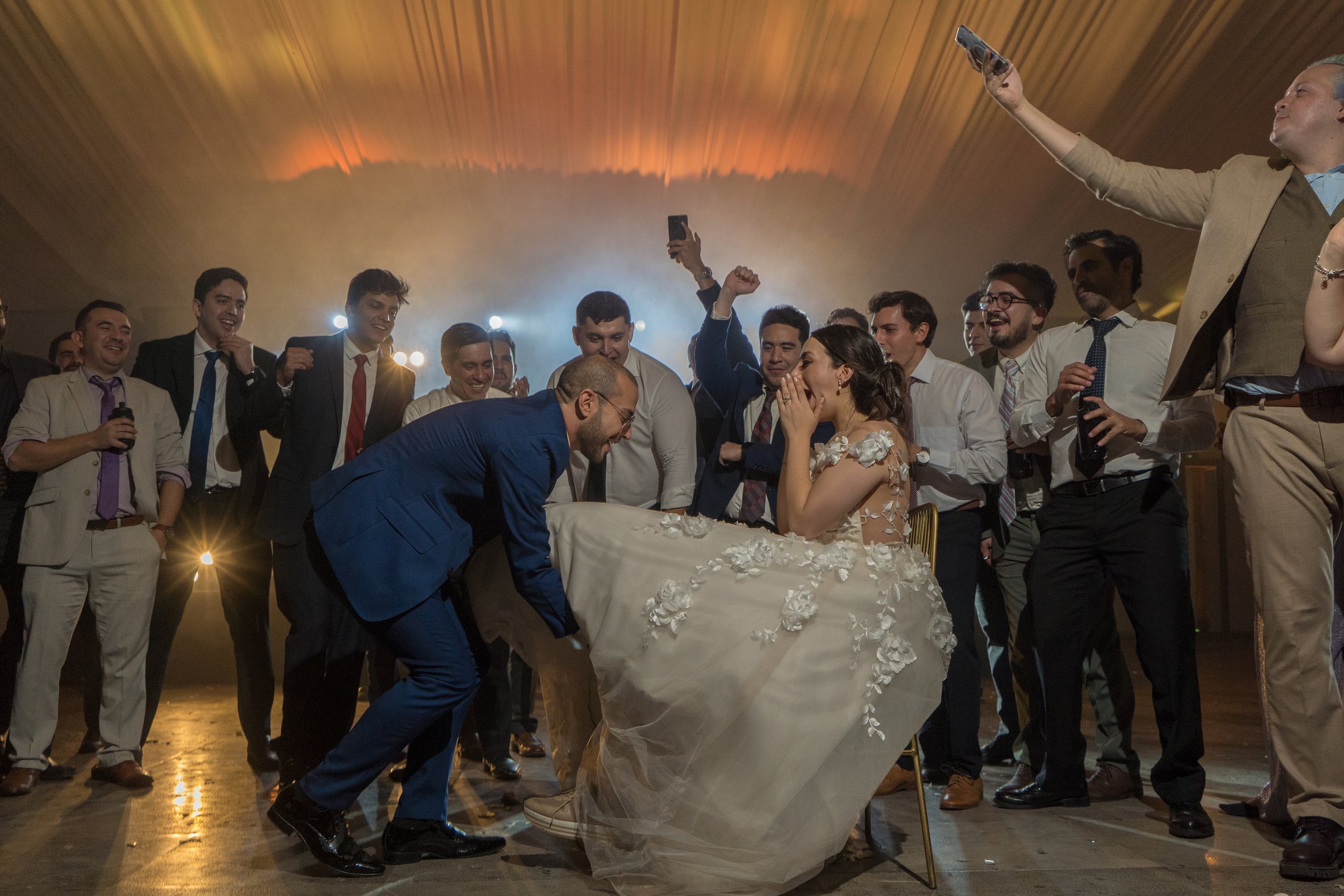 Pareja de recién casados en la celebración de su boda. La novia está sentada con su vestido de flores, cubriéndose la boca de la emoción, mientras el novio está inclinado hacia ella. Familia y amigos los rodean, riendo y tomando fotos, en un ambiente