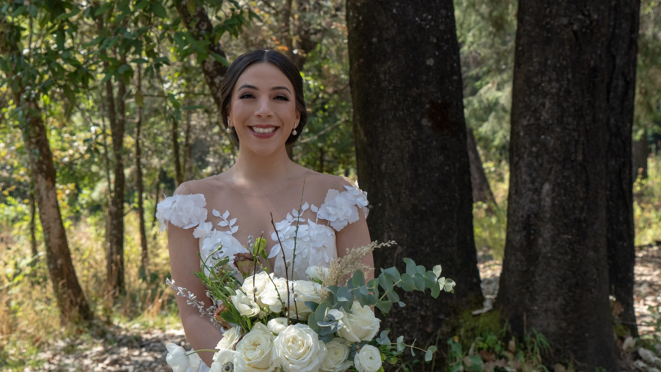 Mujer sonriente con vestido de novia sosteniendo un ramo de flores blancas, en un bosque.