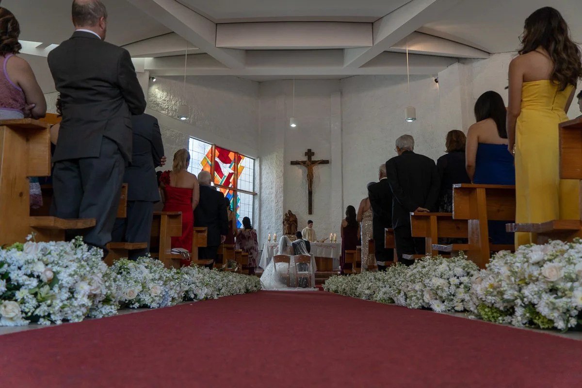 A group of people gathered inside a church during a wedding ceremony, facing an altar with a crucifix and a colorful stained glass window in the background. There are flower arrangements lining the aisle.