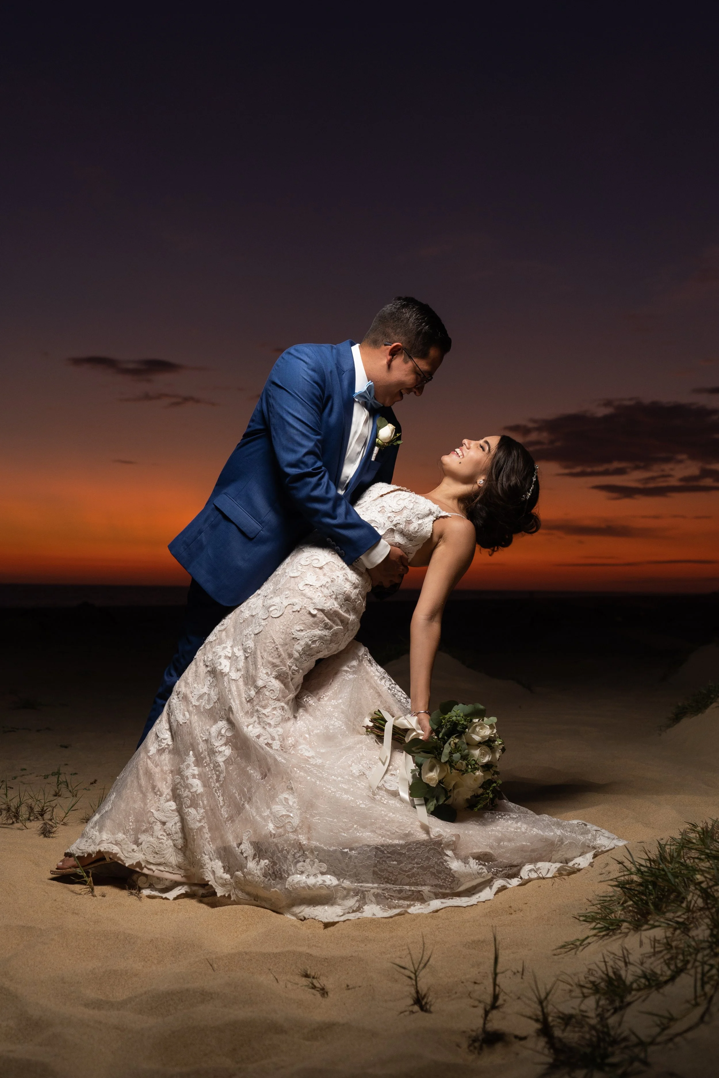 Pareja de recién casados en una playa durante el atardecer, el hombre en traje azul y la mujer con vestido de novia en una posición de danza o romántica, sosteniéndose y sonriendo.