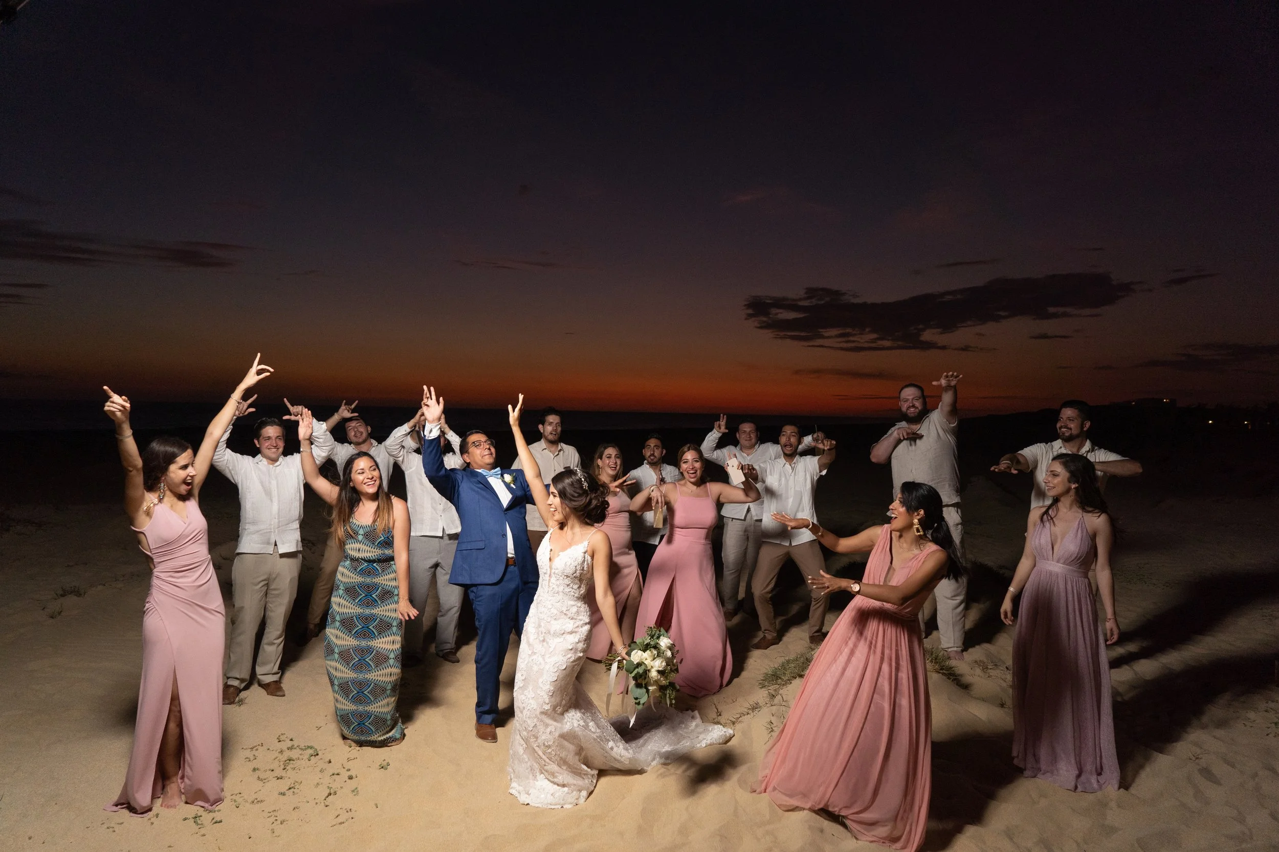 Grupo de personas en la playa durante el atardecer, celebrando una boda con disfraces elegantes y alegría.