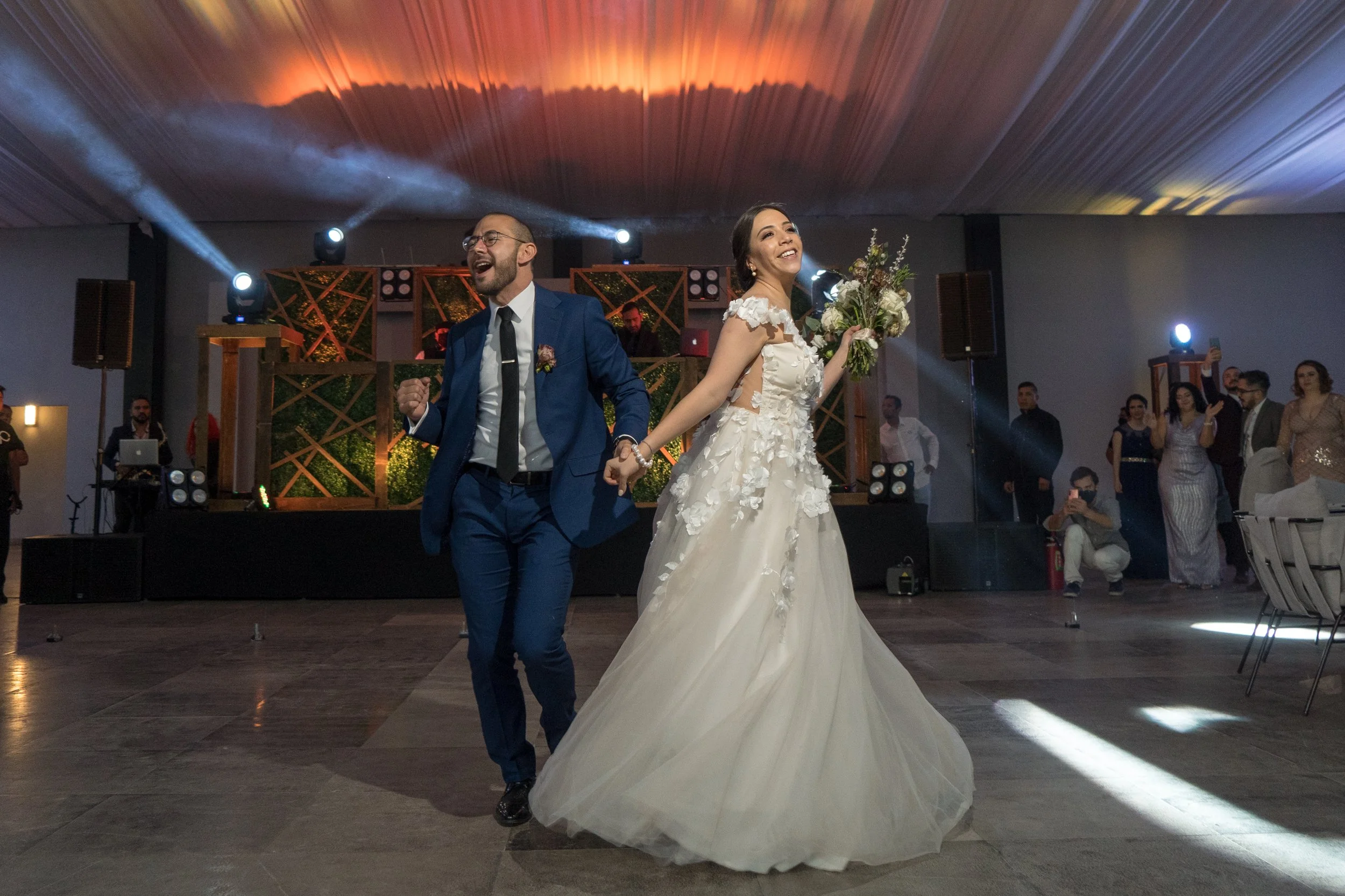 Pareja de recién casados bailando en una recepción con fondo decorativo y otros invitados observando.