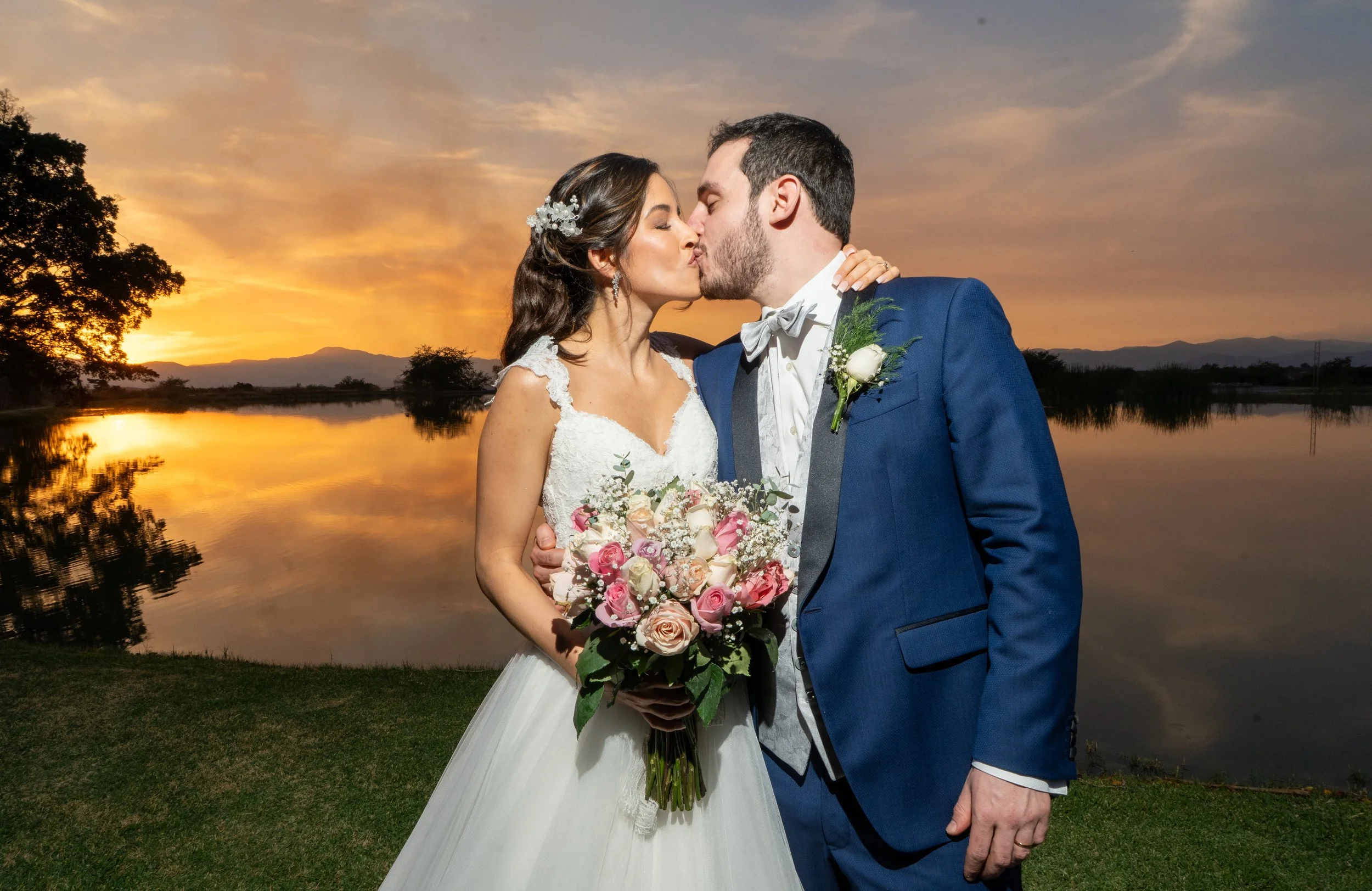 Pareja de recién casados besándose al atardecer junto a un río, con árboles y montañas en el fondo