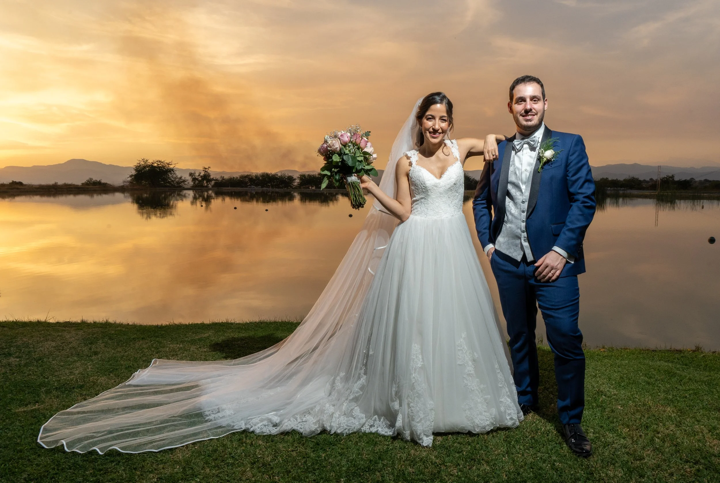 Una pareja de novios en su boda, posando frente a un lago al atardecer, con un cielo nublado y montañas al fondo. La novia viste un vestido blanco largo y la novia un traje azul con pajarita.