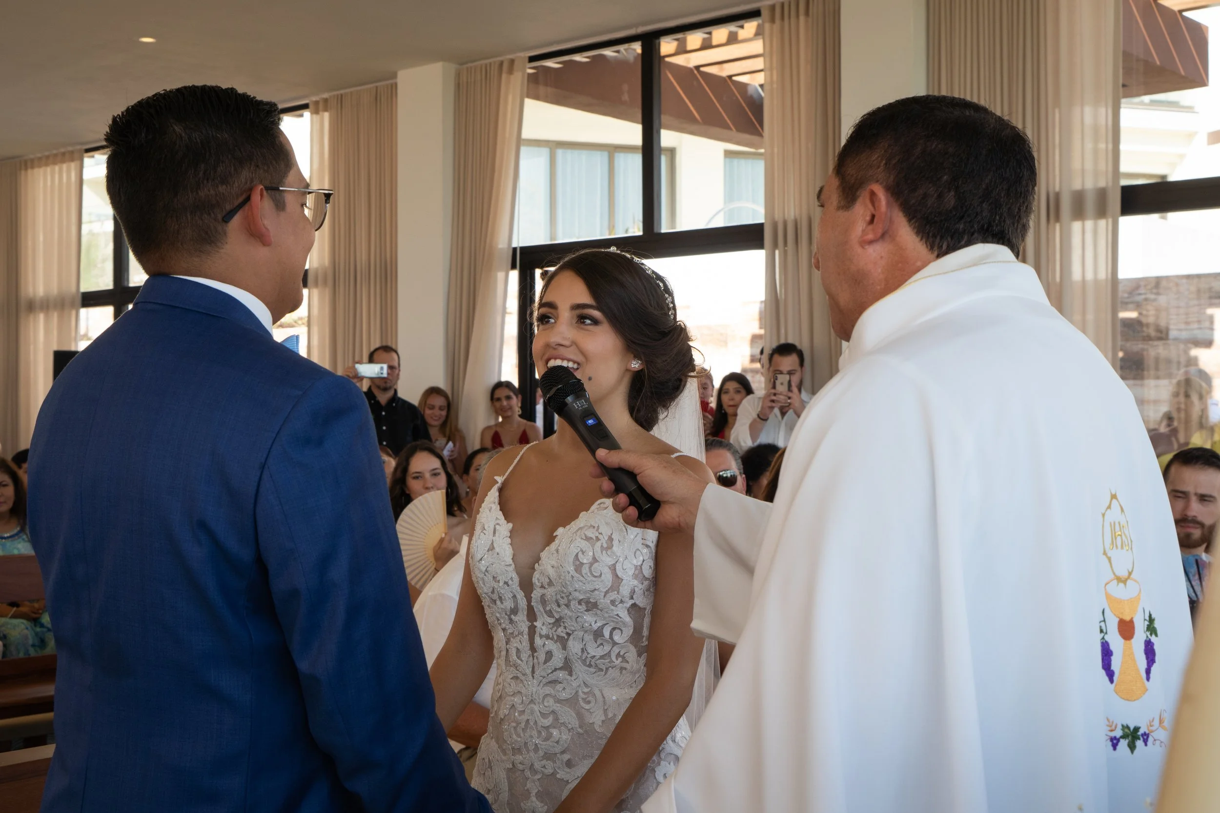 Una pareja casándose en una ceremonia religiosa, la novia sostiene las manos del novio mientras el sacerdote los bendice frente a una congregación. La novia viste un vestido de encaje blanco y el novio un saco azul. La ceremonia se realiza en un luga