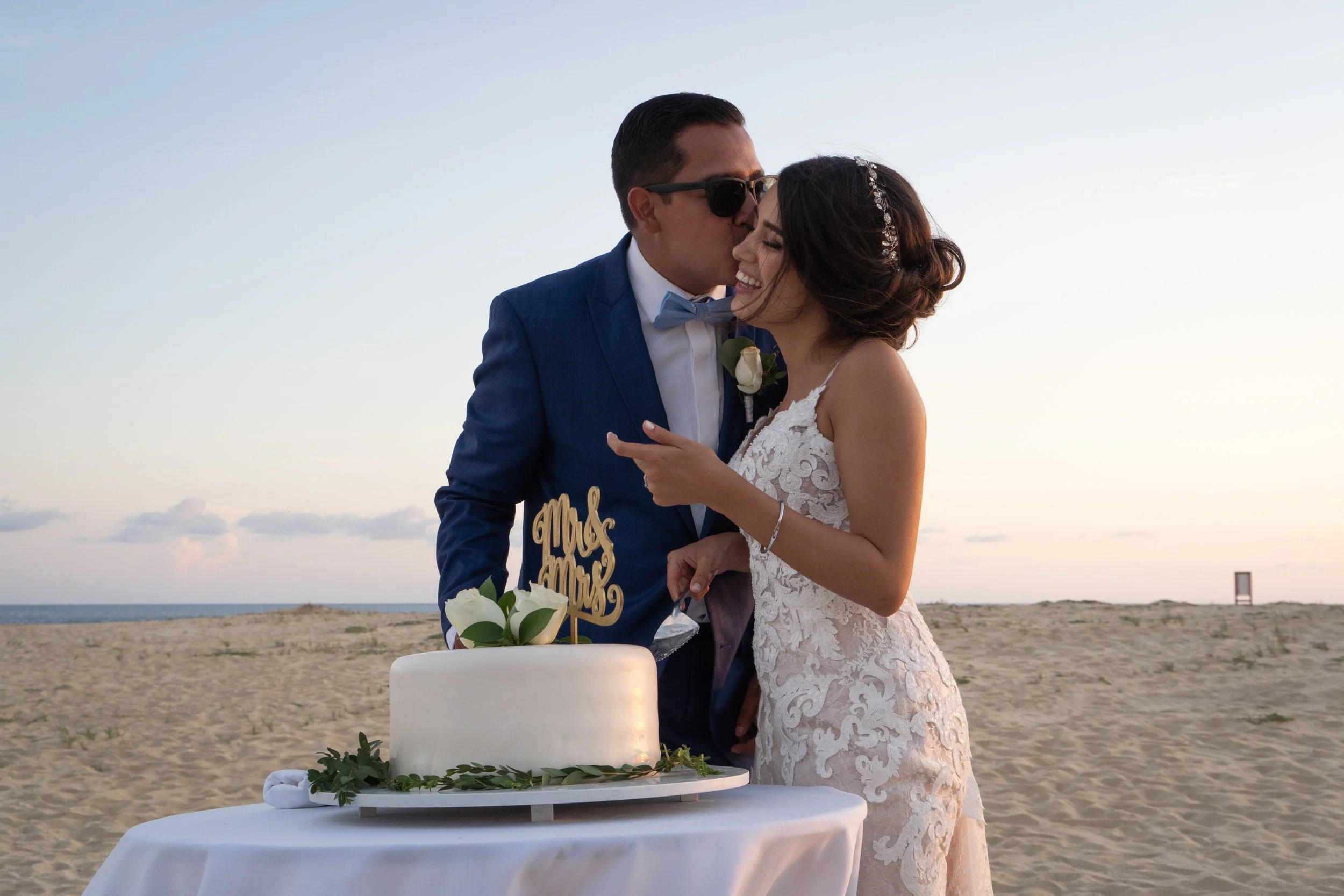 Una pareja de recién casados en la playa compartiendo un momento de felicidad junto a una tarta de bodas decorada con flores blancas y una señal que dice 'Mr & Mrs'.