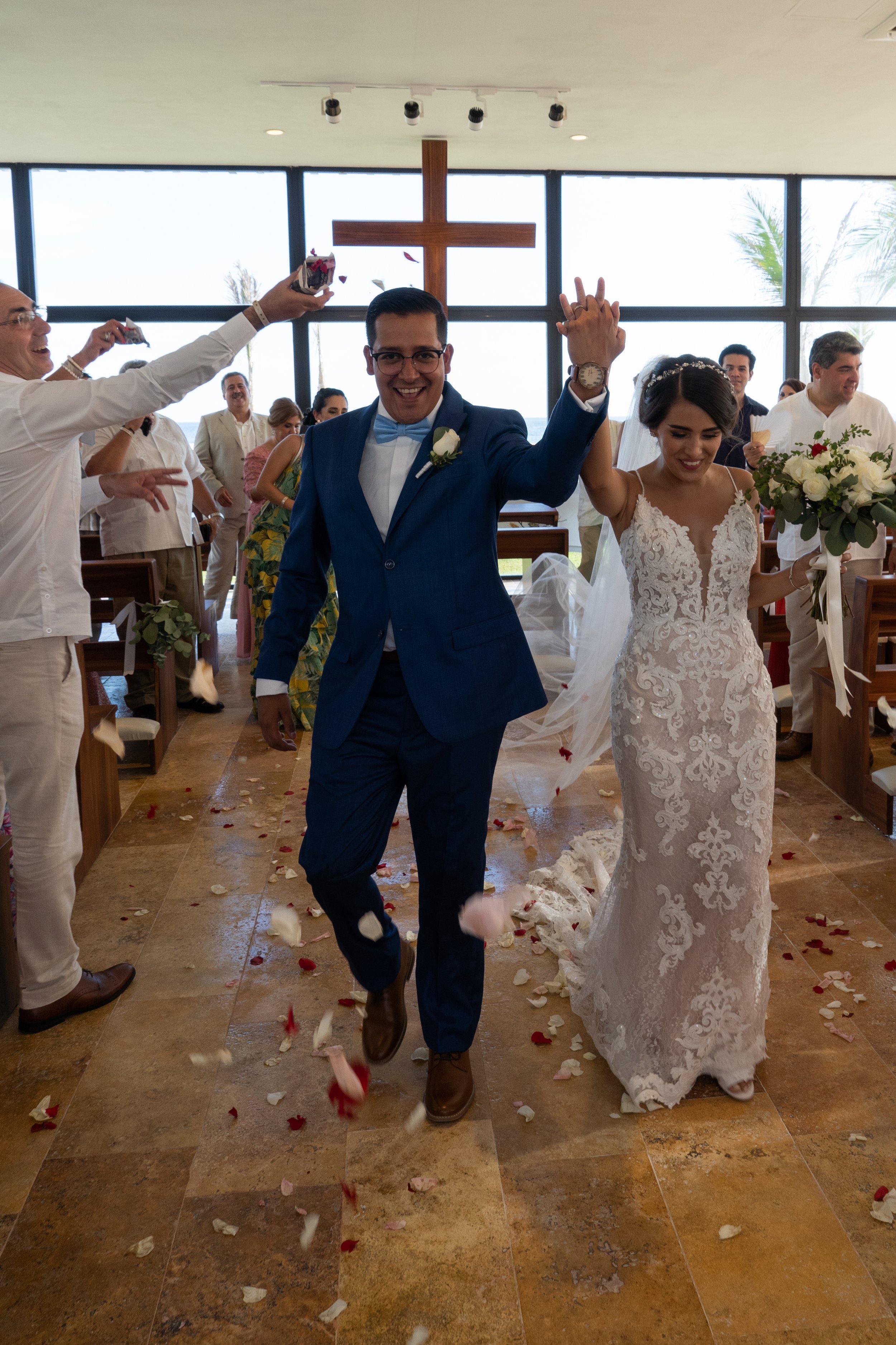 Pareja de recién casados celebrando en iglesia, el hombre lleva traje azul y la mujer vestido de novia con ramo, rodeados de invitados y confeti en el suelo.