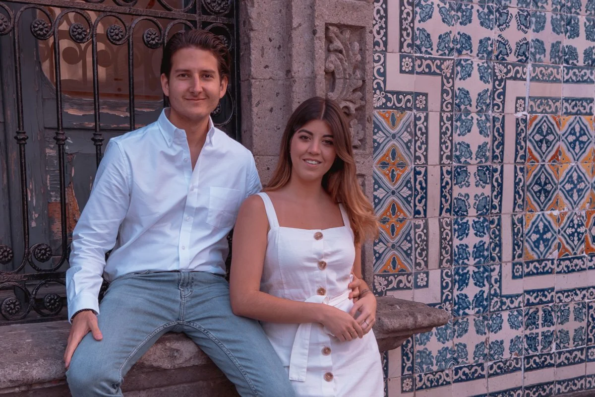 A young man and woman standing in front of a decorative tiled wall. The man is wearing a white shirt and gray jeans, while the woman is wearing a white dress with buttons. They are smiling at the camera.